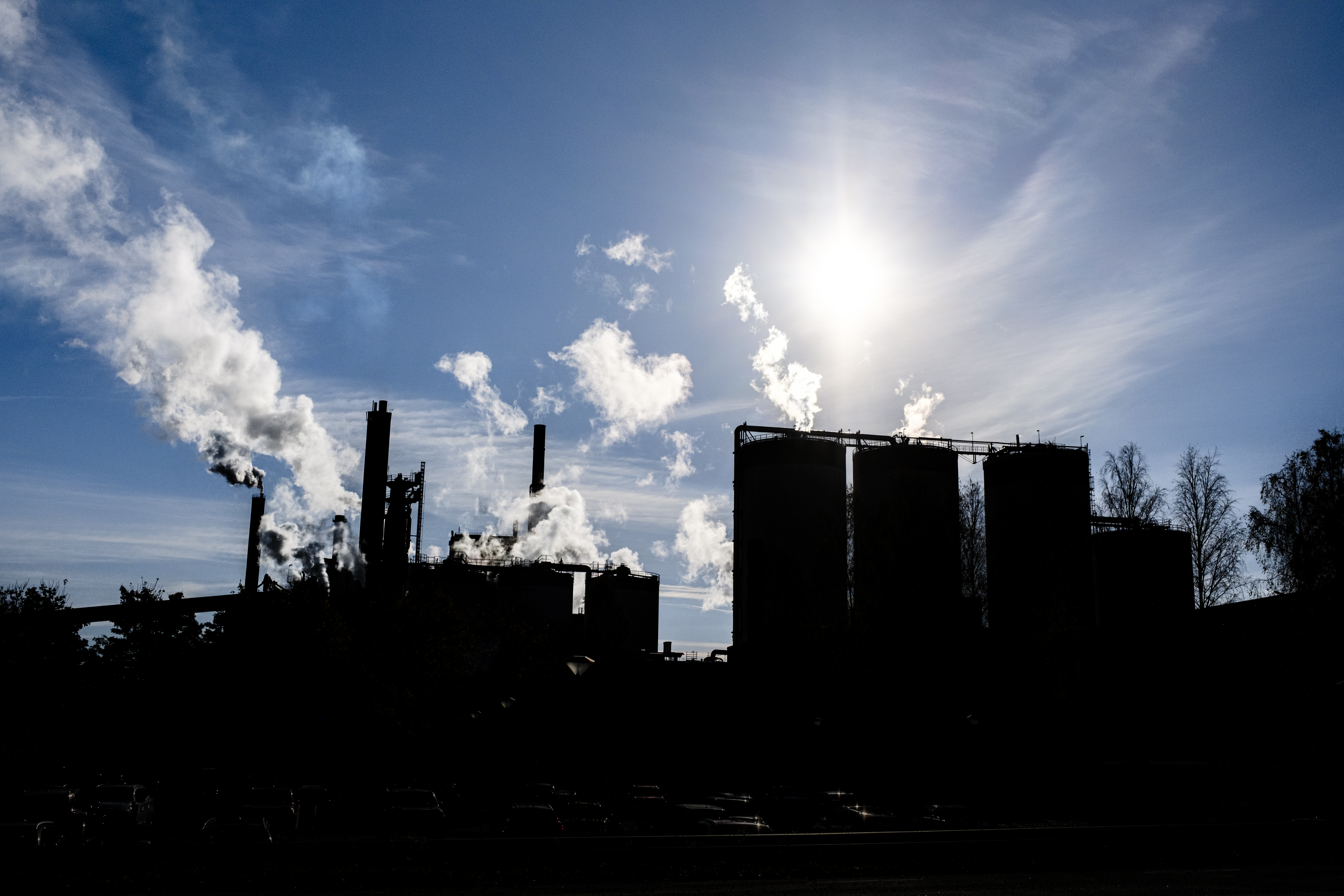 En fabrik med flera skorstenar som släpper ut vit rök under en sol och en delvis molnig himmel.