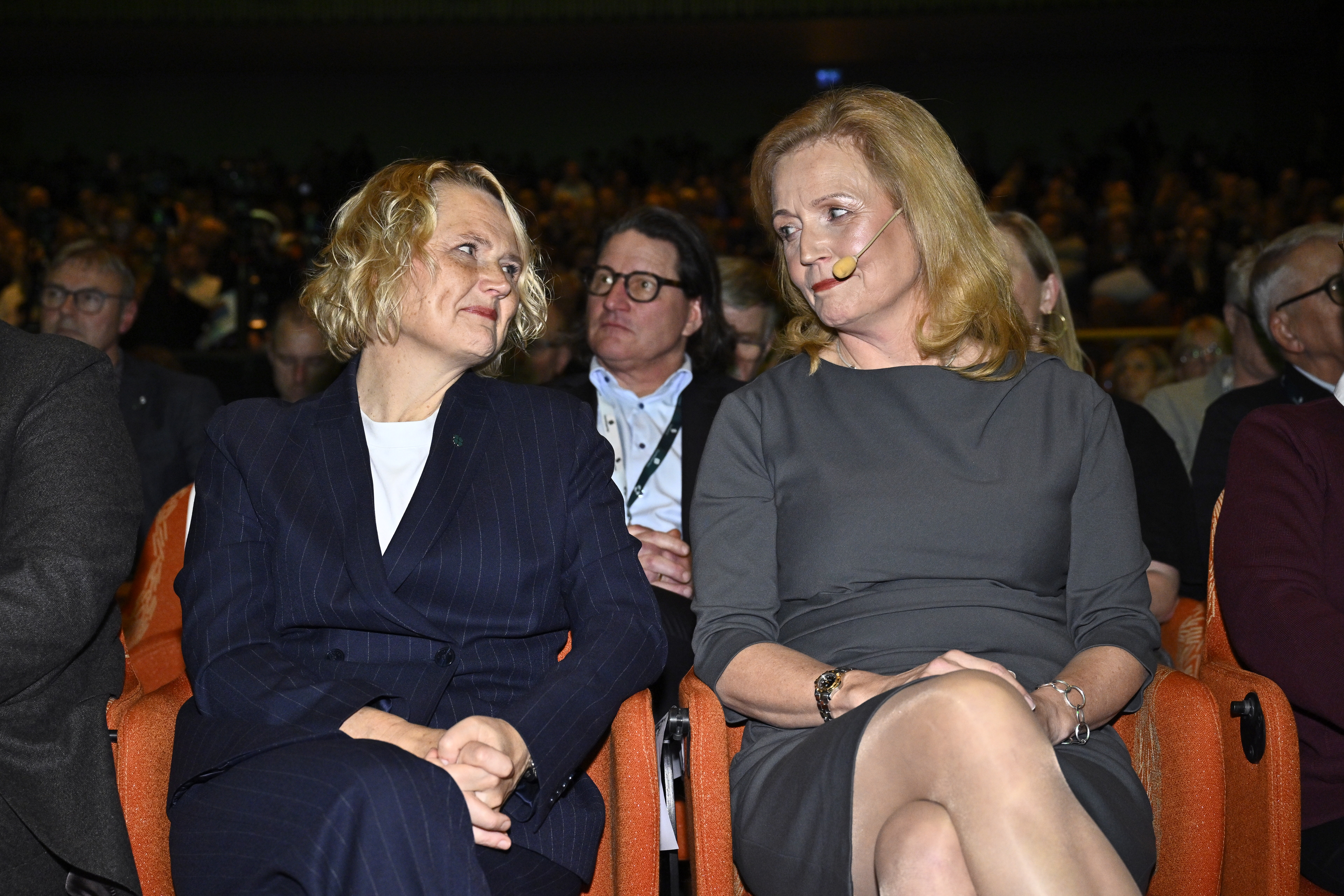 Två kvinnor i affärskläder sitter och samtalar på ett evenemang med mikrofoner. Publik sitter i rader bakom dem.