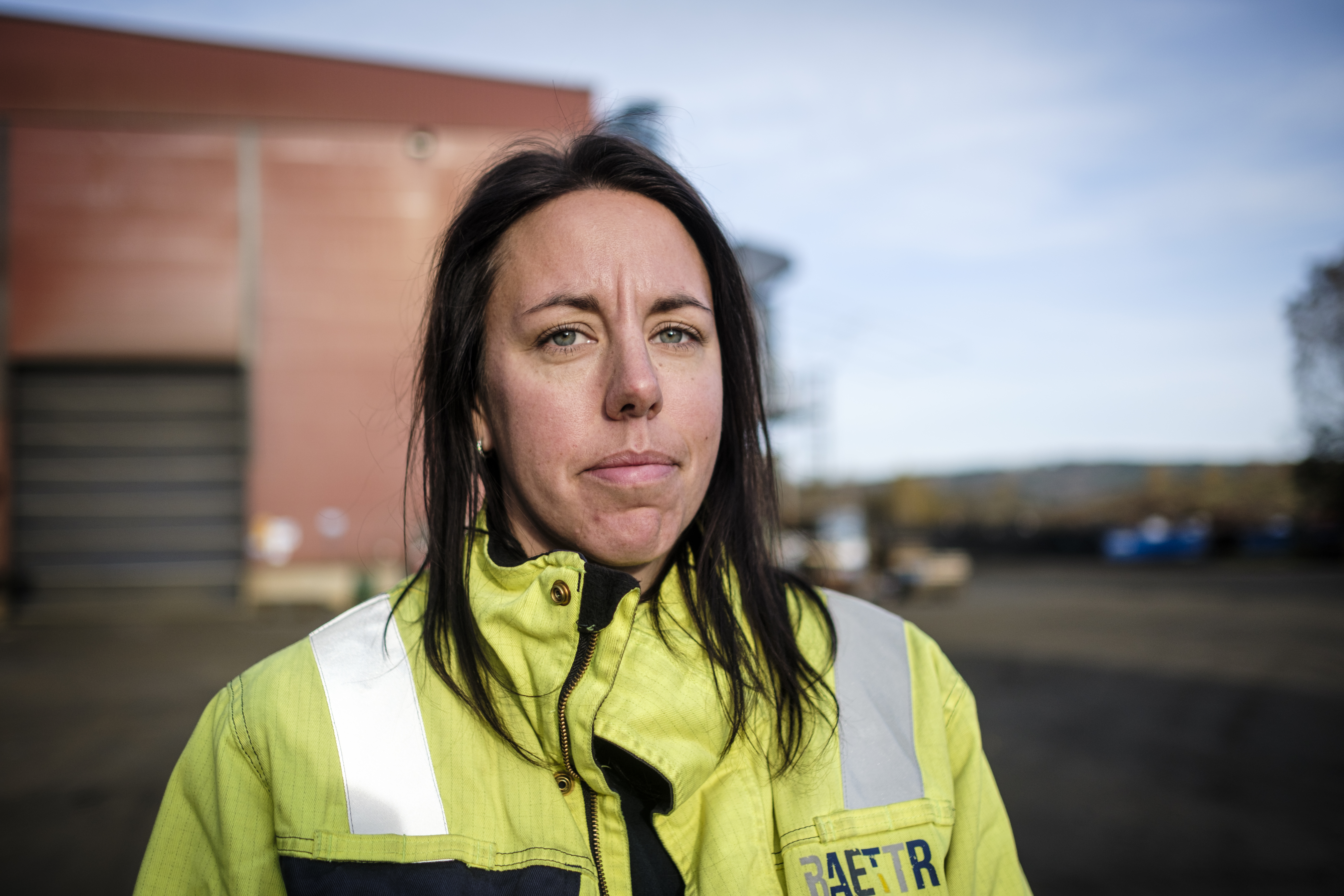 En person med varseljacka står utomhus i ett industriområde, med en stor byggnad och utrustning i bakgrunden.