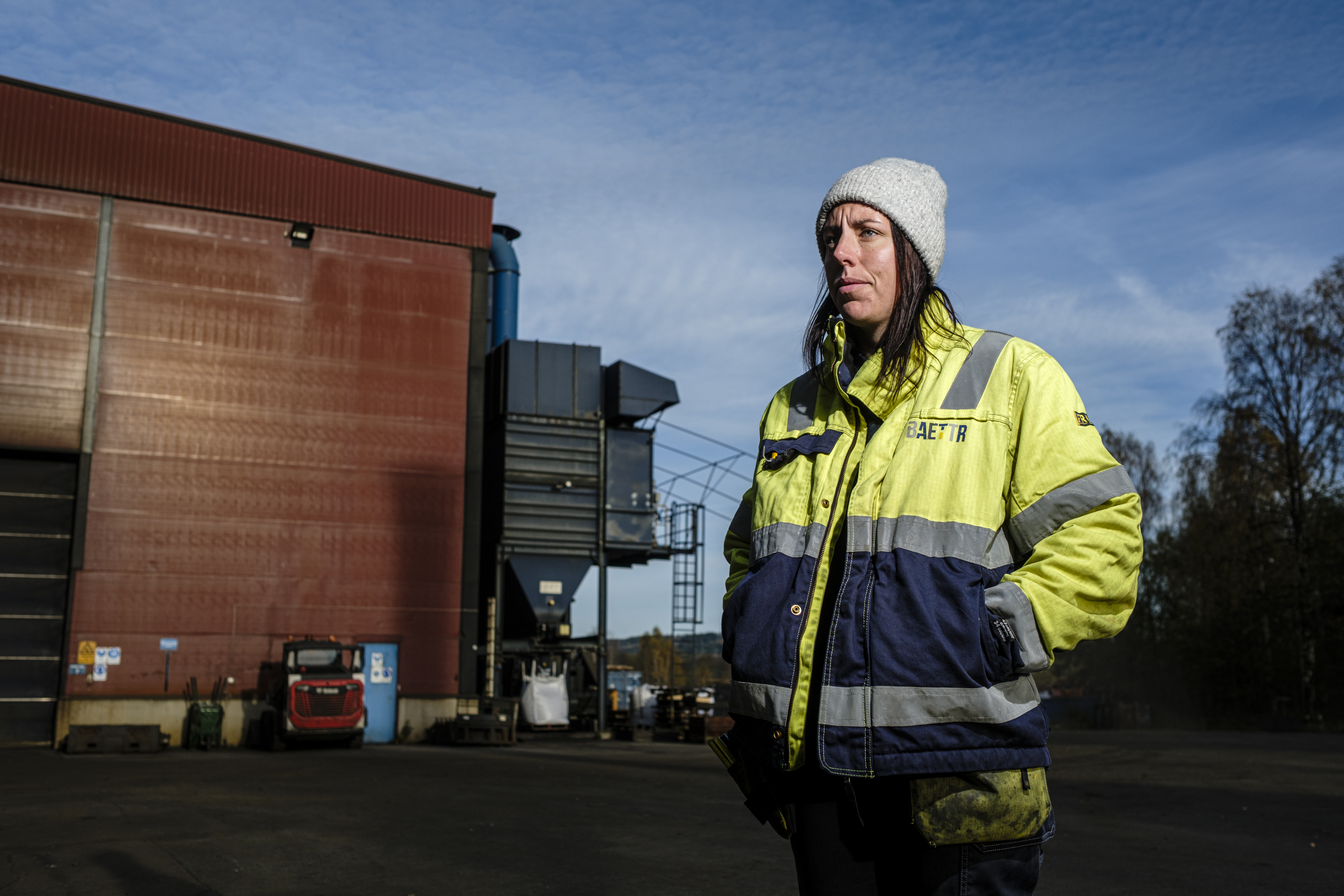 Anna Berglund med grå mössa och varseljacka står utomhus framför en industribyggnad under en klar dag.