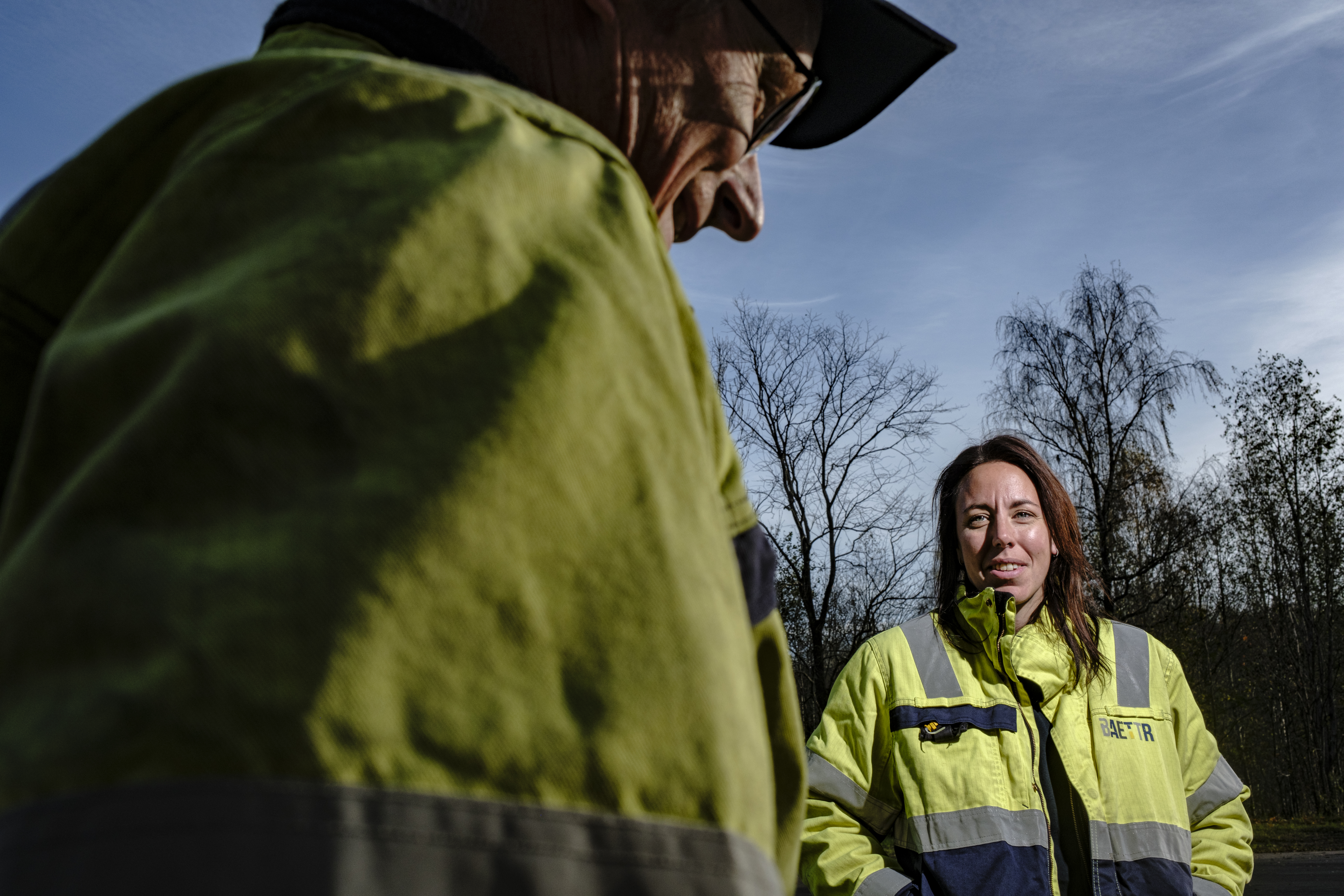 Andreas Ericsson och Anna Berglund med varseljackor står utomhus under en blå himmel, med träd utan löv i bakgrunden.