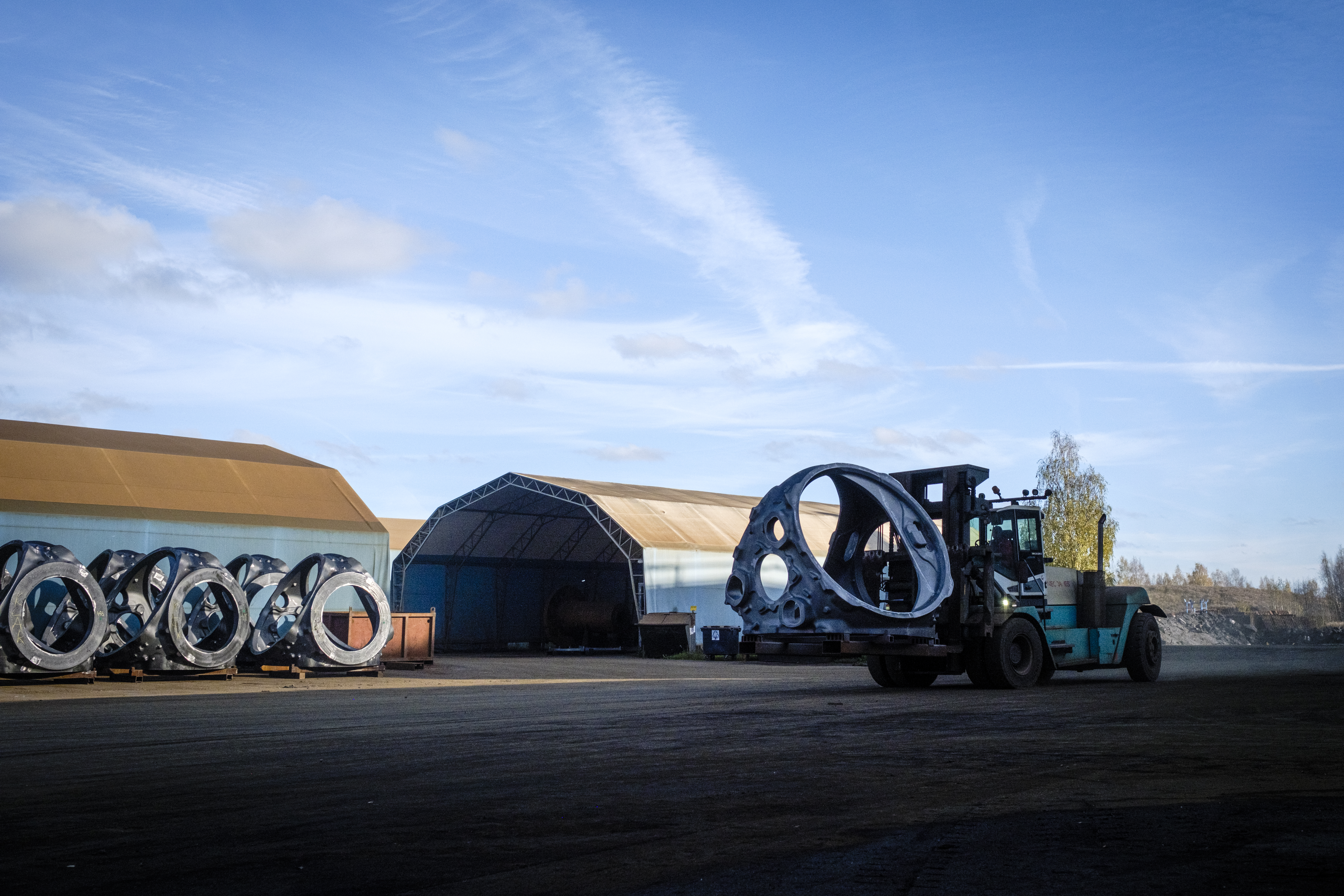 En truck flyttar ett stort metallnav på en industrigård med flera lagerbyggnader och fler metallringar staplade intill.