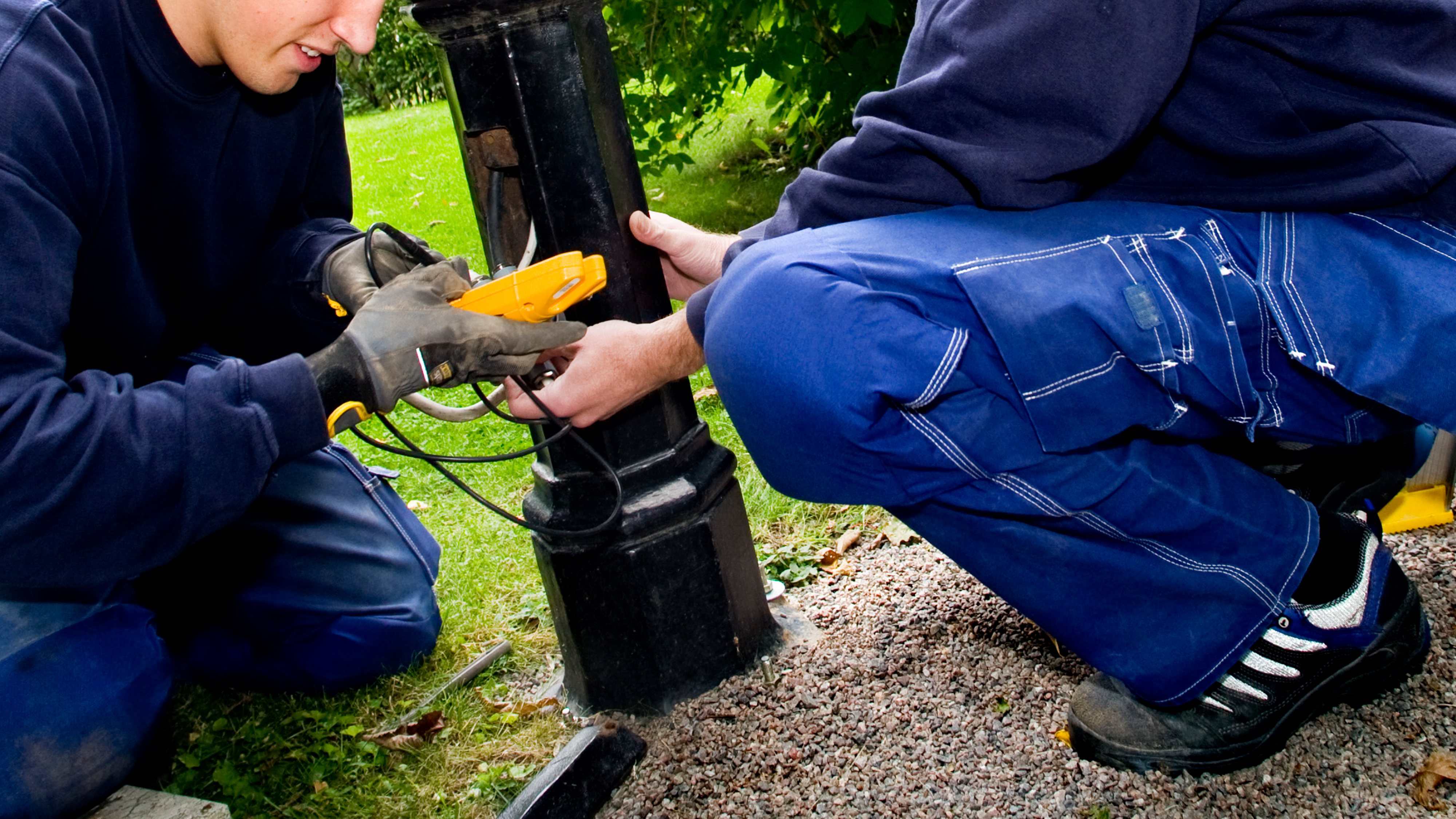 Två arbetare i blå arbetskläder använder en elektrisk testapparat på en svart stolpe i grus med gräs och träd i bakgrunden.
