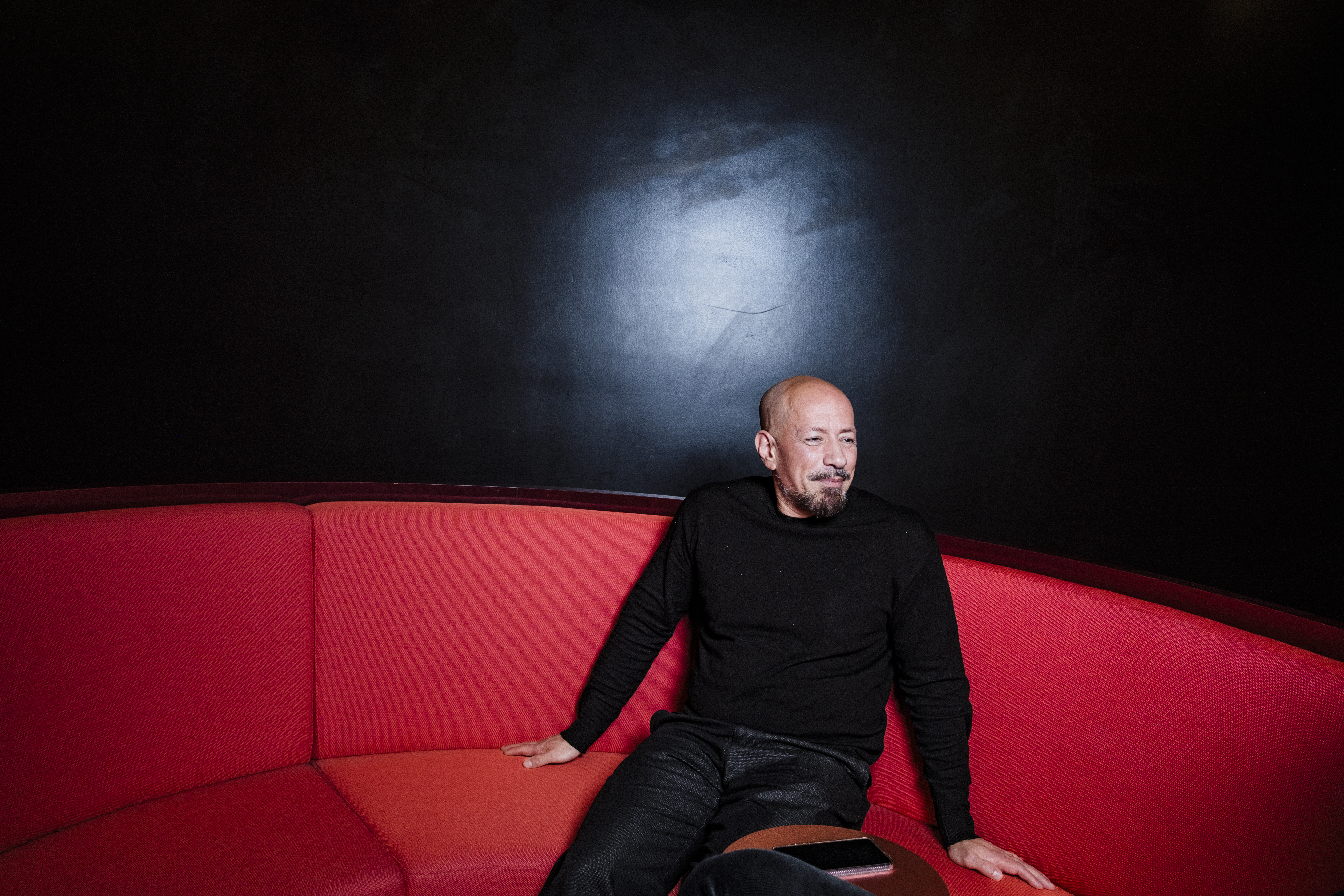 En man i svart tröja sitter på en rundad röd soffa mot mörk bakgrund, med en smartphone bredvid sig.