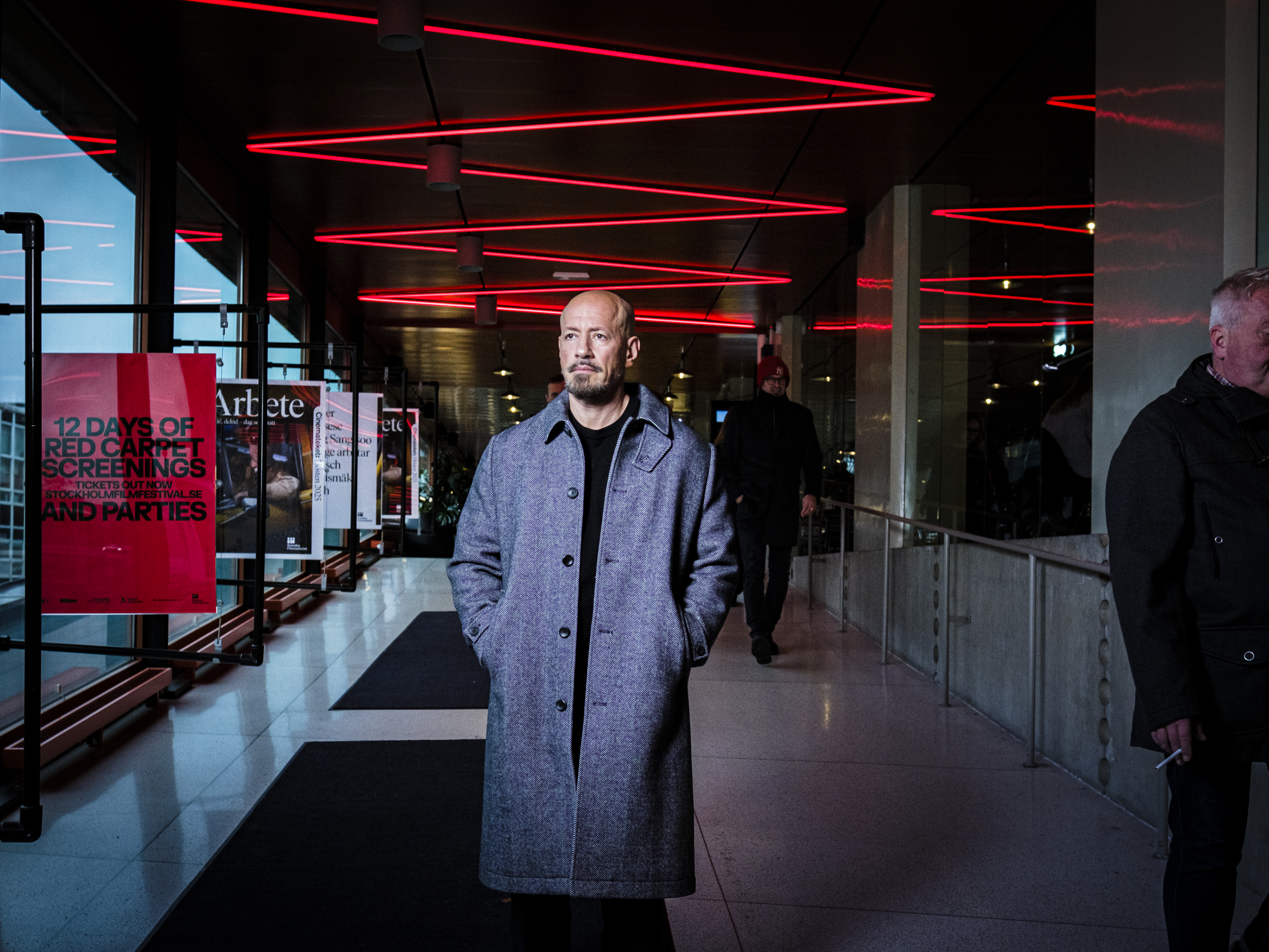 En man i grå rock står inomhus under röda neonljus med händerna i fickorna. Folk syns i bakgrunden vid glasväggar och affischer.