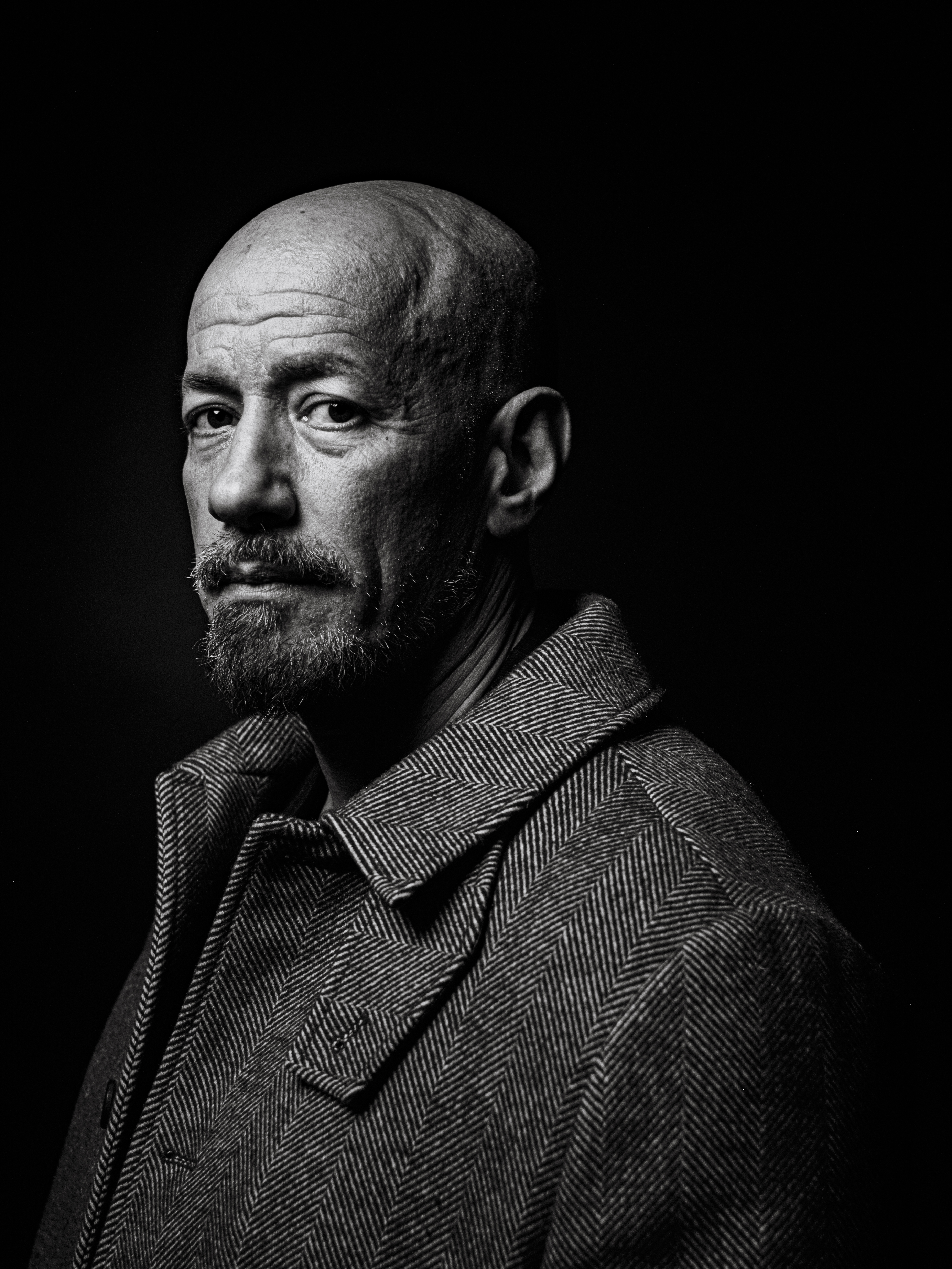 Svarthvitt porträtt av medelålders man med rakat huvud, skägg och mustasch, i kappa, tittande snett åt sidan mot mörk bakgrund.