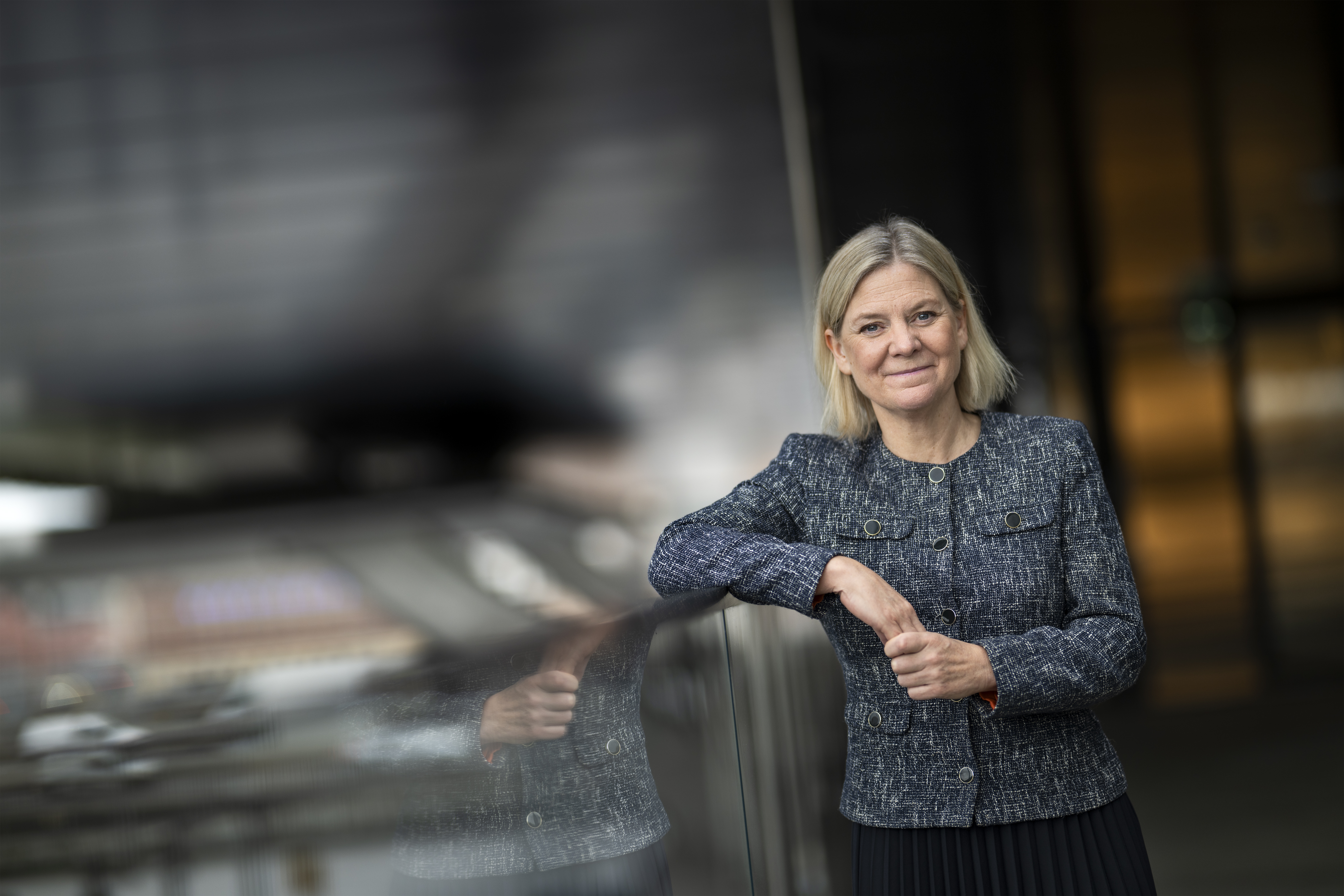 En kvinna med blont hår i blå jacka står inomhus och lutar sig mot en reflekterande yta, suddig bakgrund.