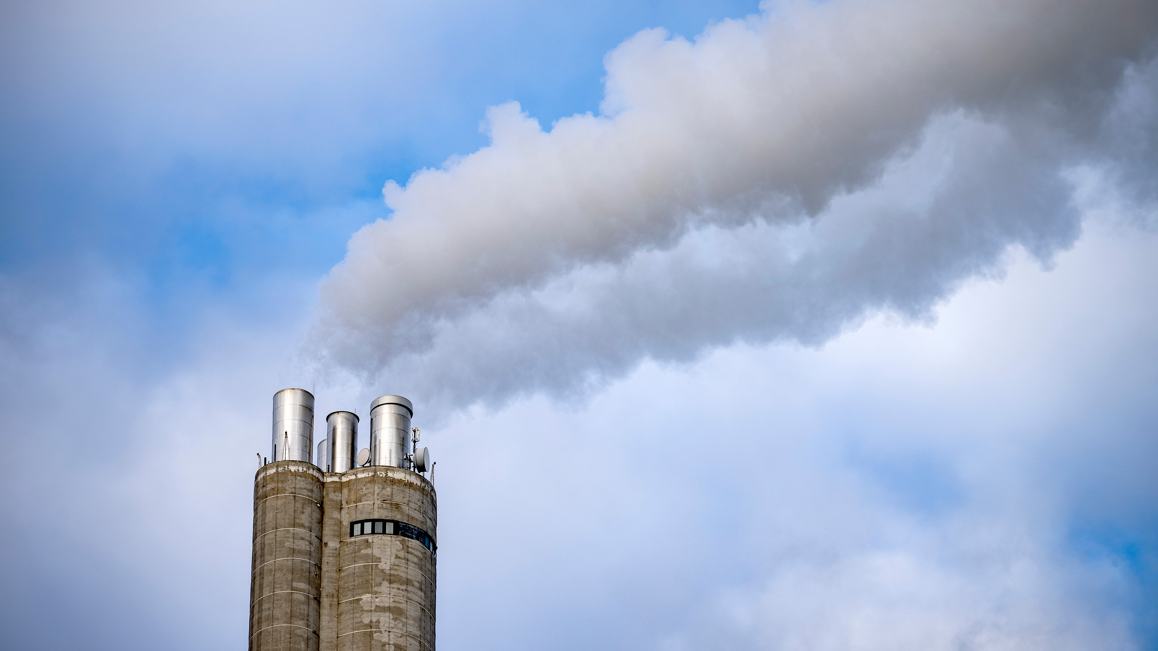 Tre stora industritorn släpper ut vit rök mot en delvis molnig blå himmel.