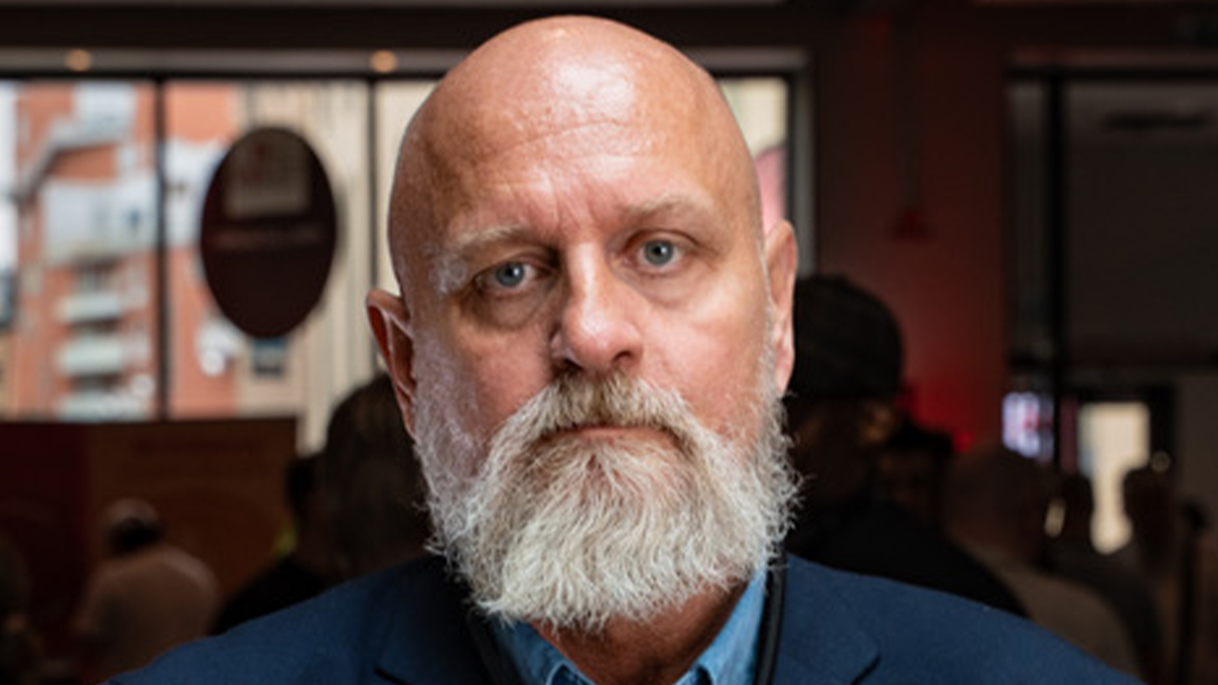 En man med vitt skägg, iförd blå jacka, står inomhus med människor och fönster i bakgrunden.