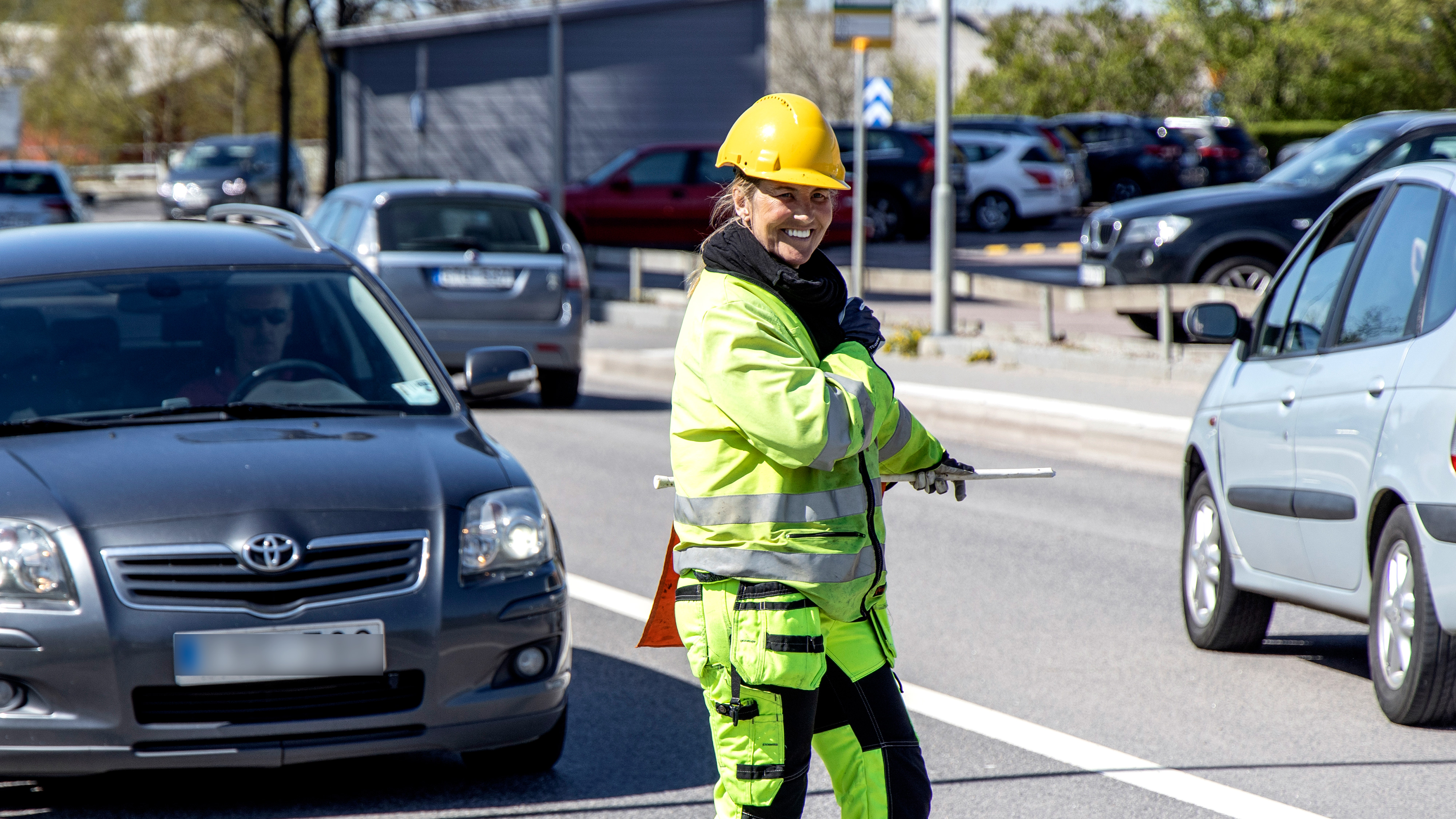 En byggarbetare med gul hjälm och varselkläder dirigerar trafiken på en gata med bilar på båda sidor.