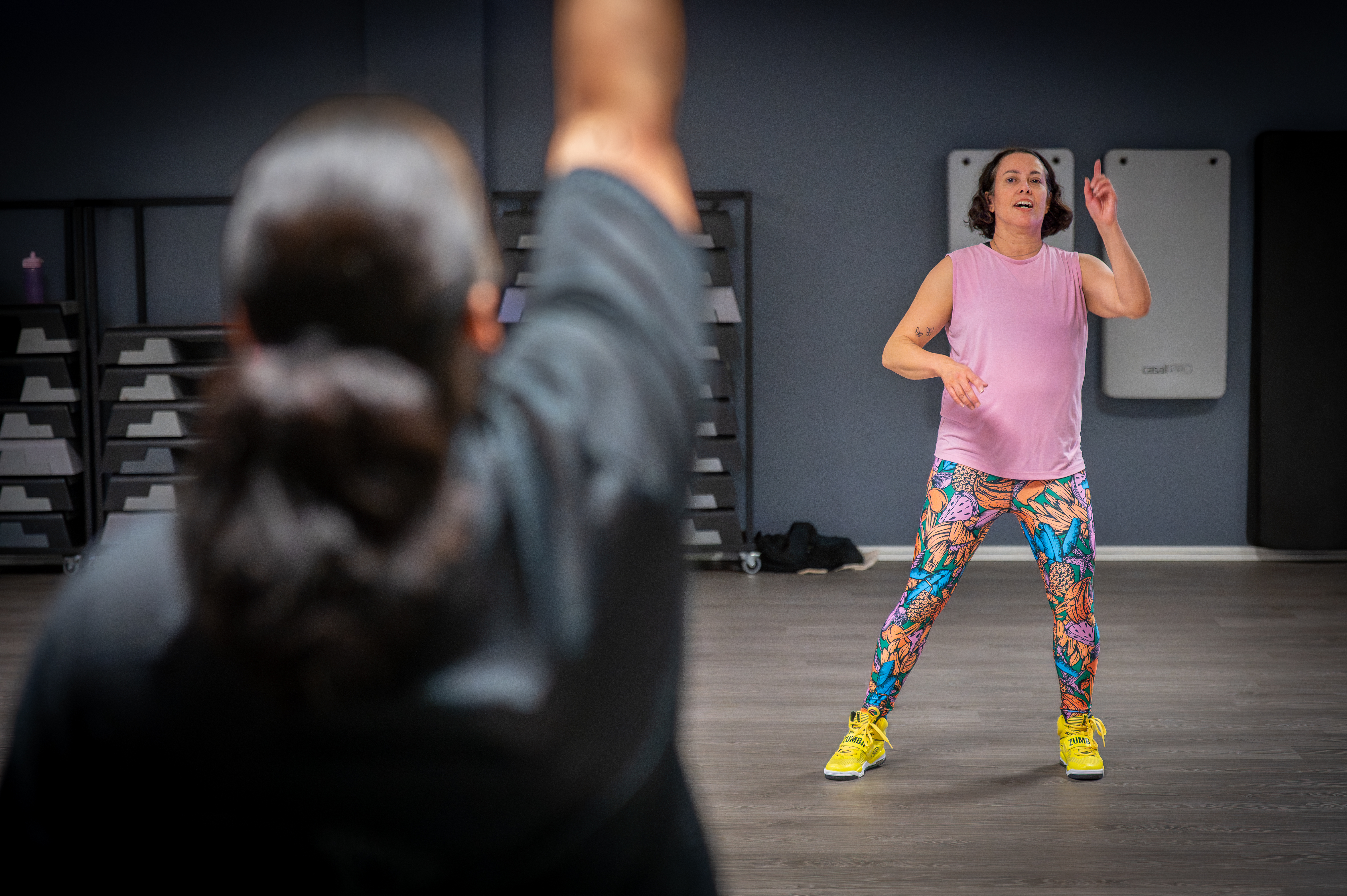 En kvinna i leggings och gula skor leder en träningsklass inomhus, medan en annan deltagare lyfter en arm i förgrunden.