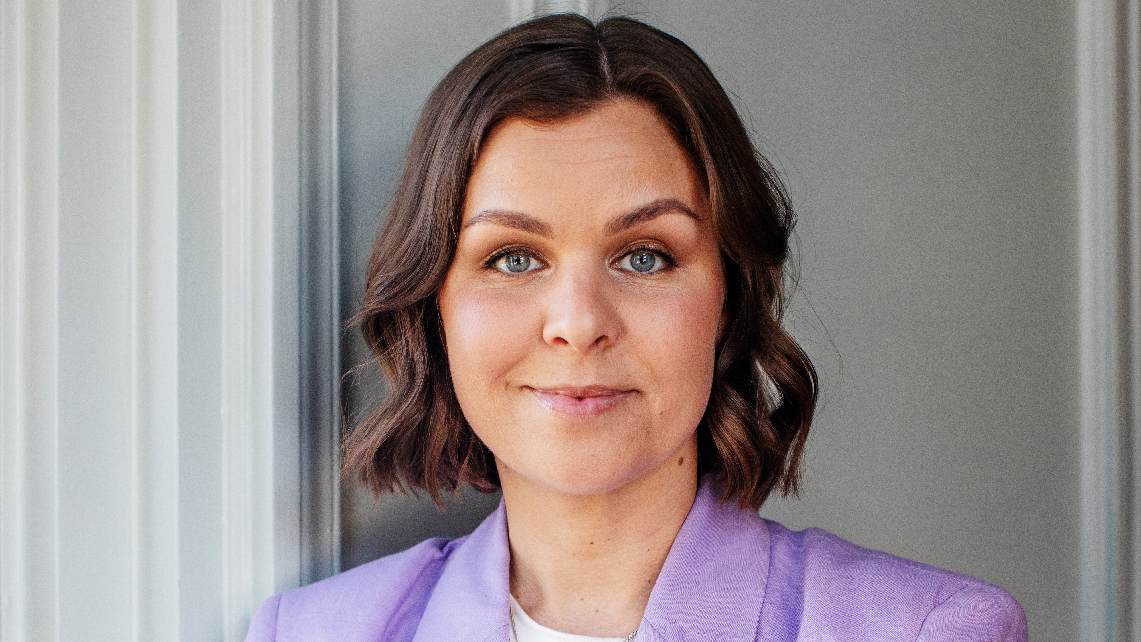 Lina Ejlertsson, med kort brunt hår, blå ögon, ljuslila jacka och vit tröja, står framför en gråvit vägg.