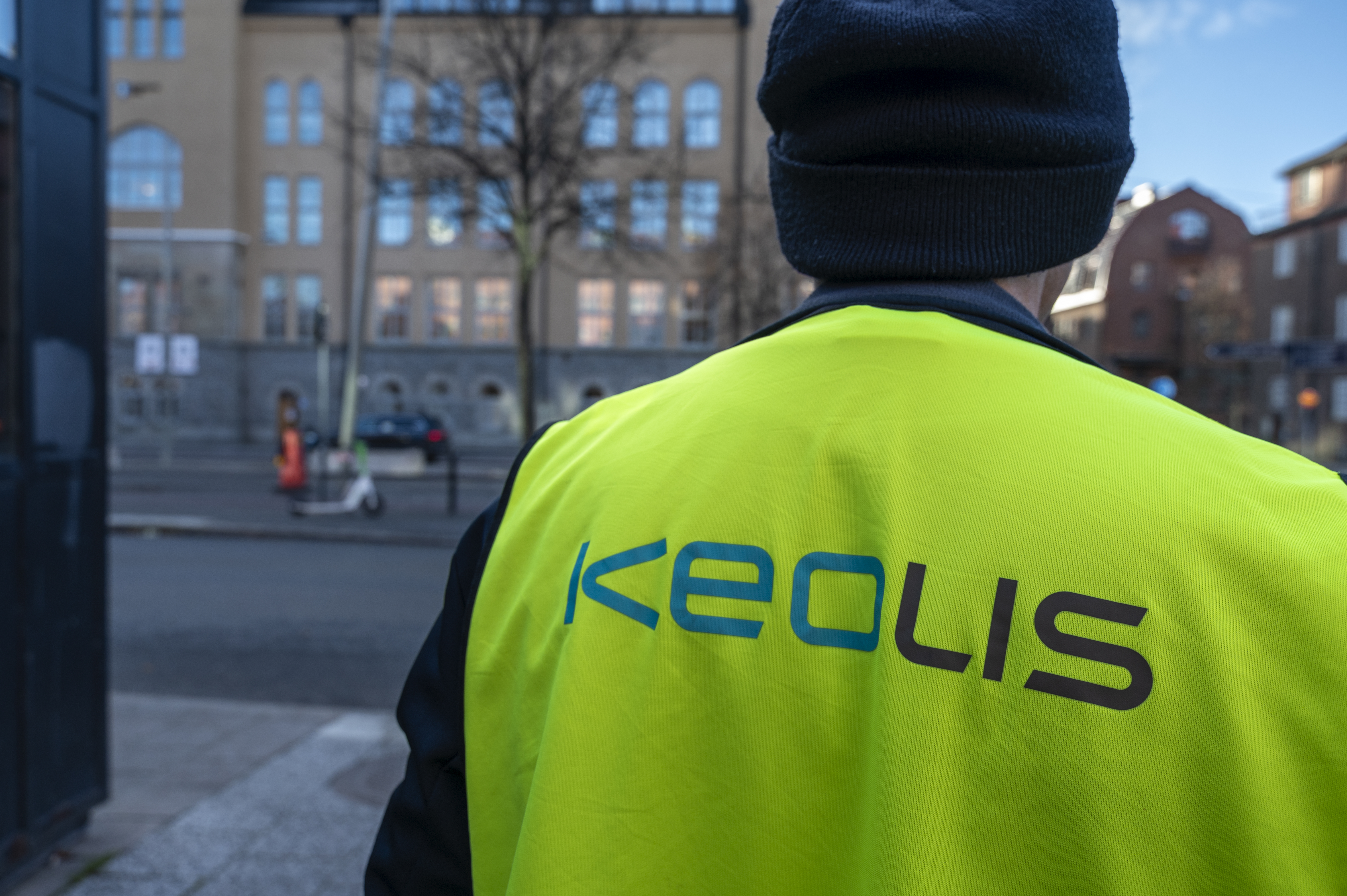 En person med en gul väst med "Keolis"-logotyp står på en stadsgata nära byggnader.