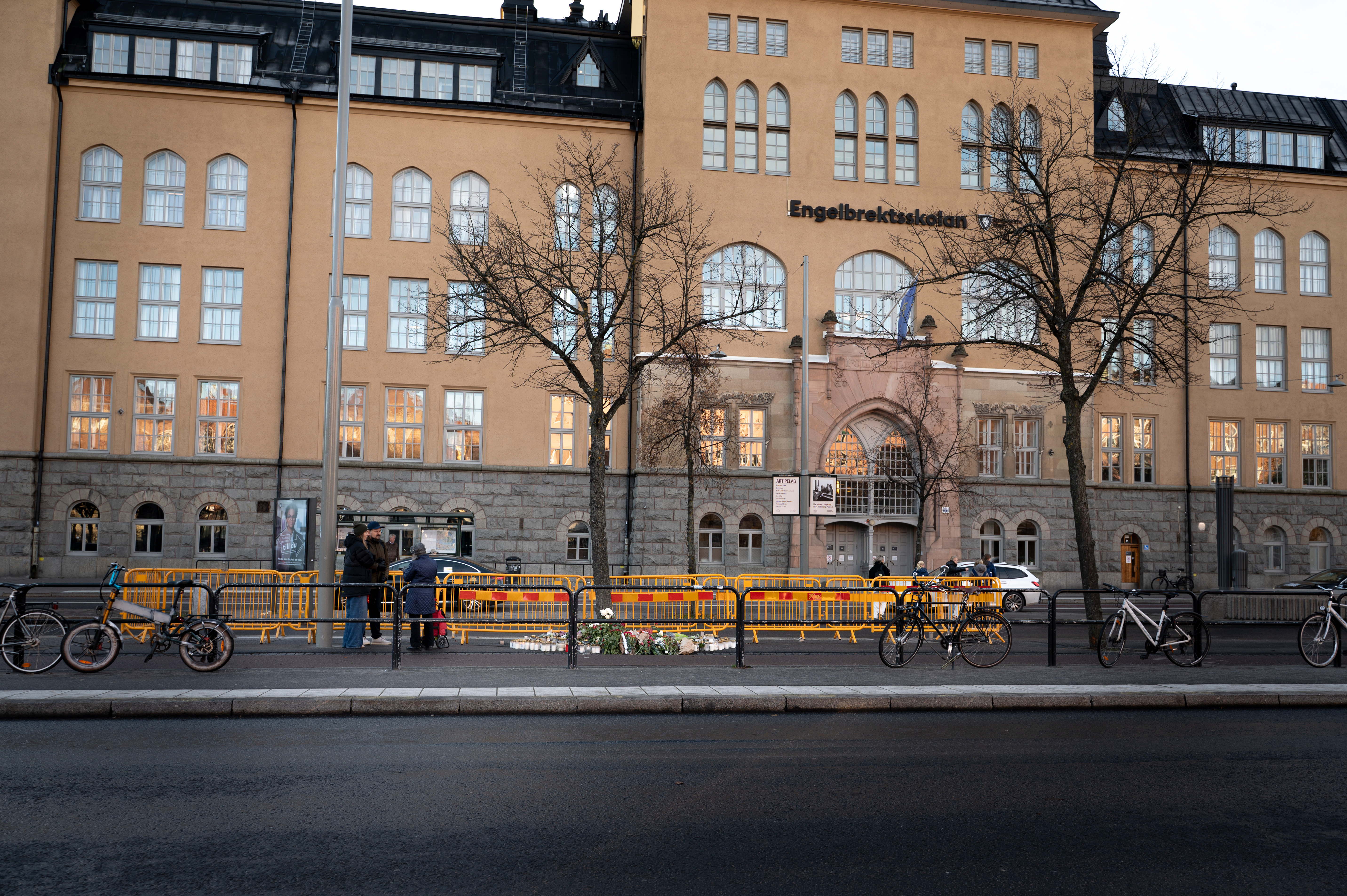 En byggnad med skylten "Engelbrektsskolan", välvda fönster, träd, cyklar och gula avspärrningar vid trottoaren.