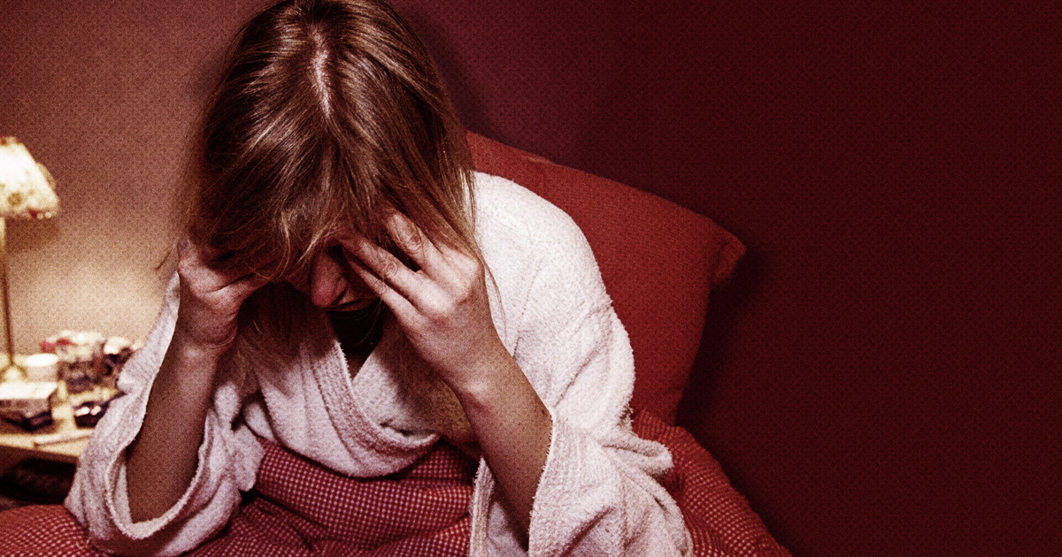 En person i vit morgonrock sitter på en säng med huvudet nedåt och håller sitt huvud med båda händerna i ett rum.