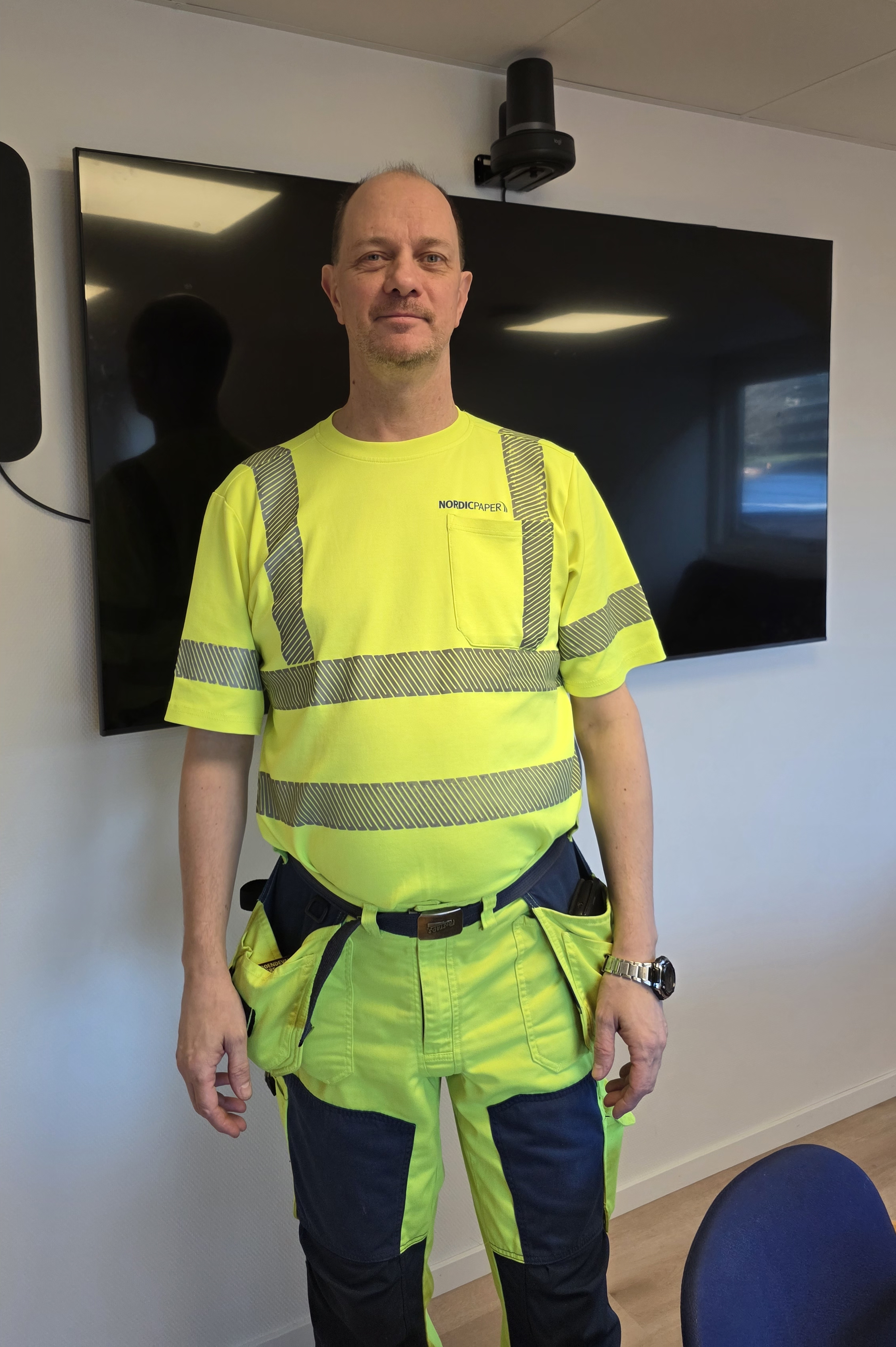 En man med en gul varselarbetsuniform står inomhus framför en stor väggmonterad TV-skärm.