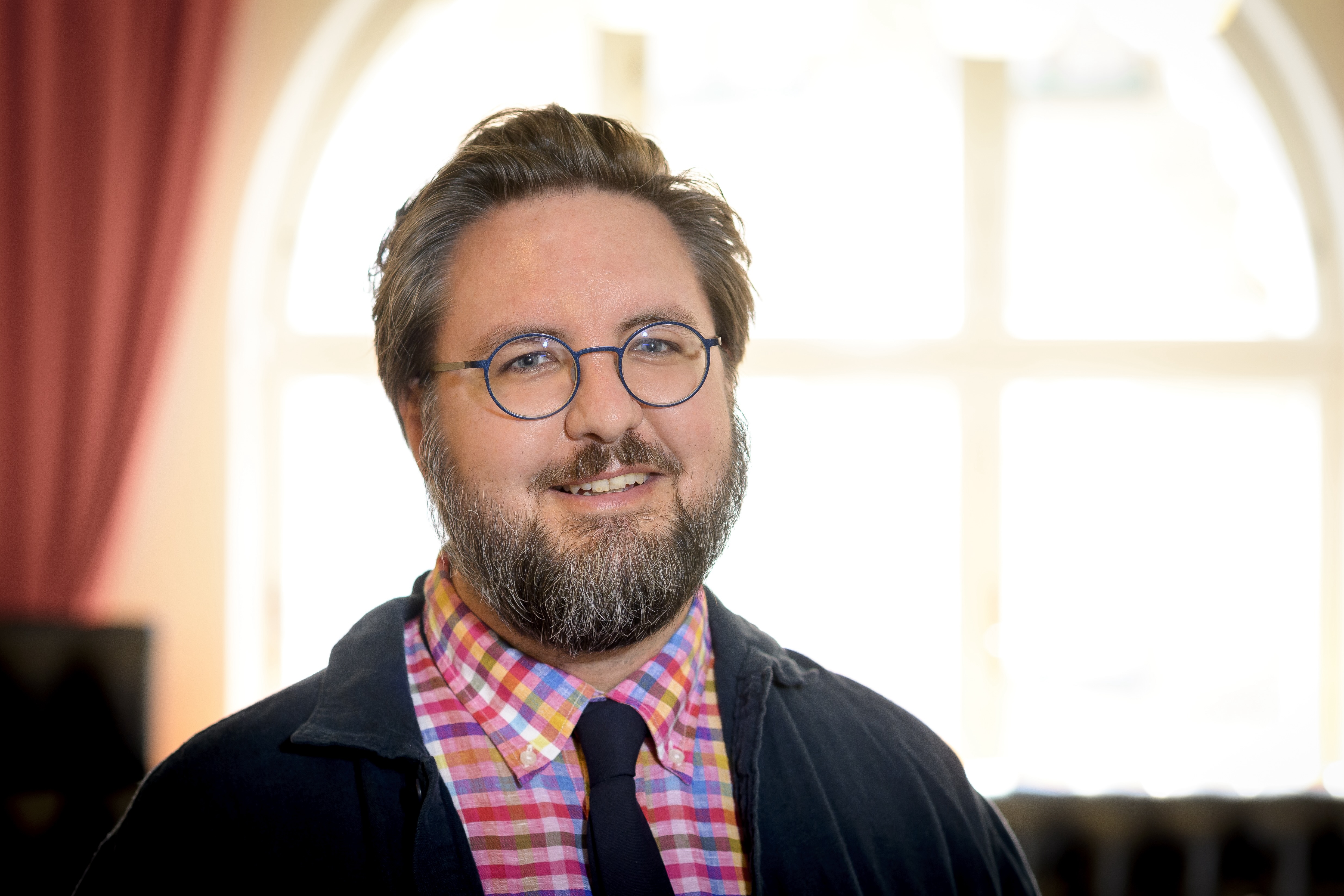 En man med glasögon och skägg, iklädd rutig skjorta, slipsar och mörk jacka står inomhus framför ett stort fönster med gardiner.