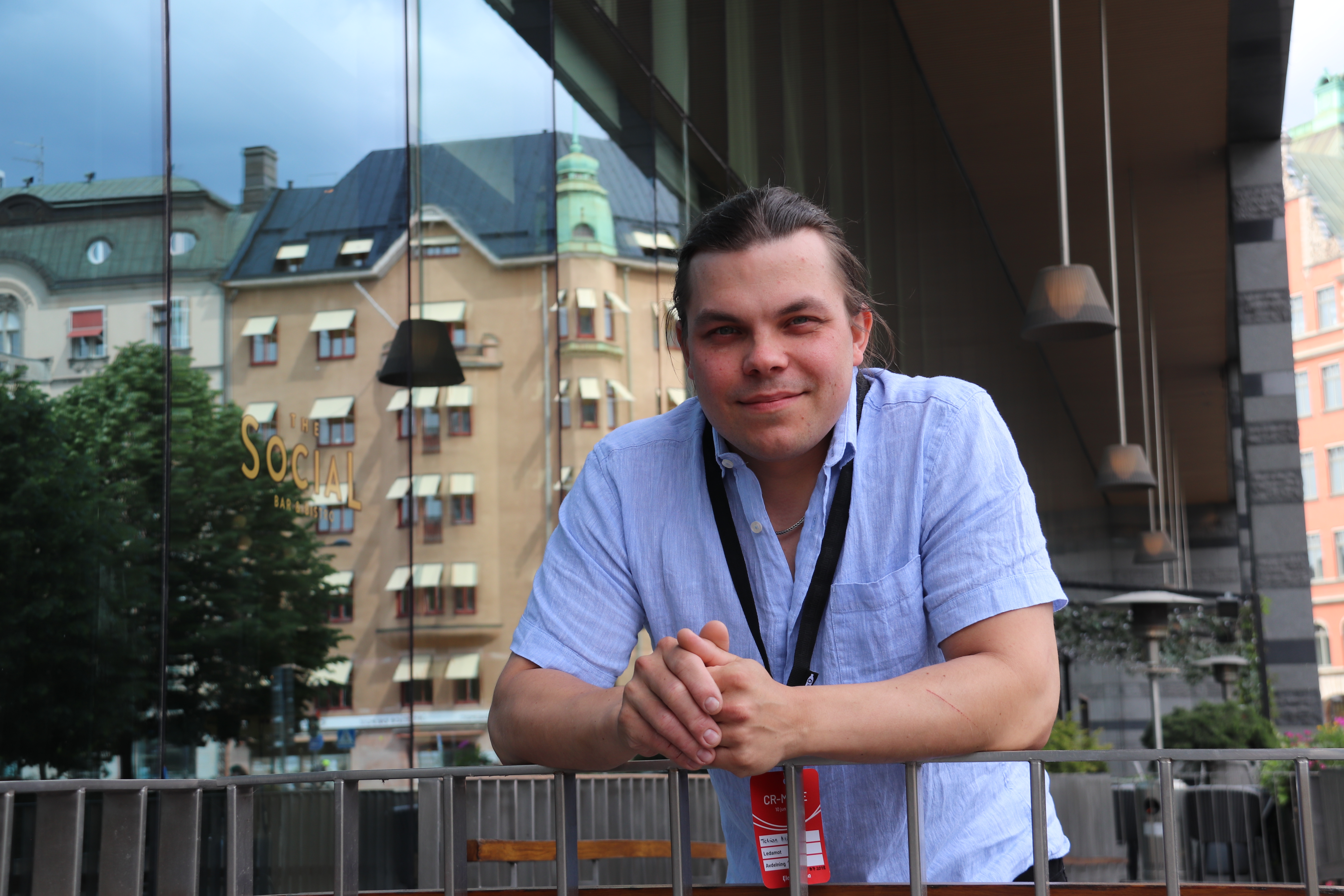 En person i en ljusblå skjorta lutar sig mot ett räcke utomhus. Stadsbyggnader och träd speglas i glasväggen bakom.