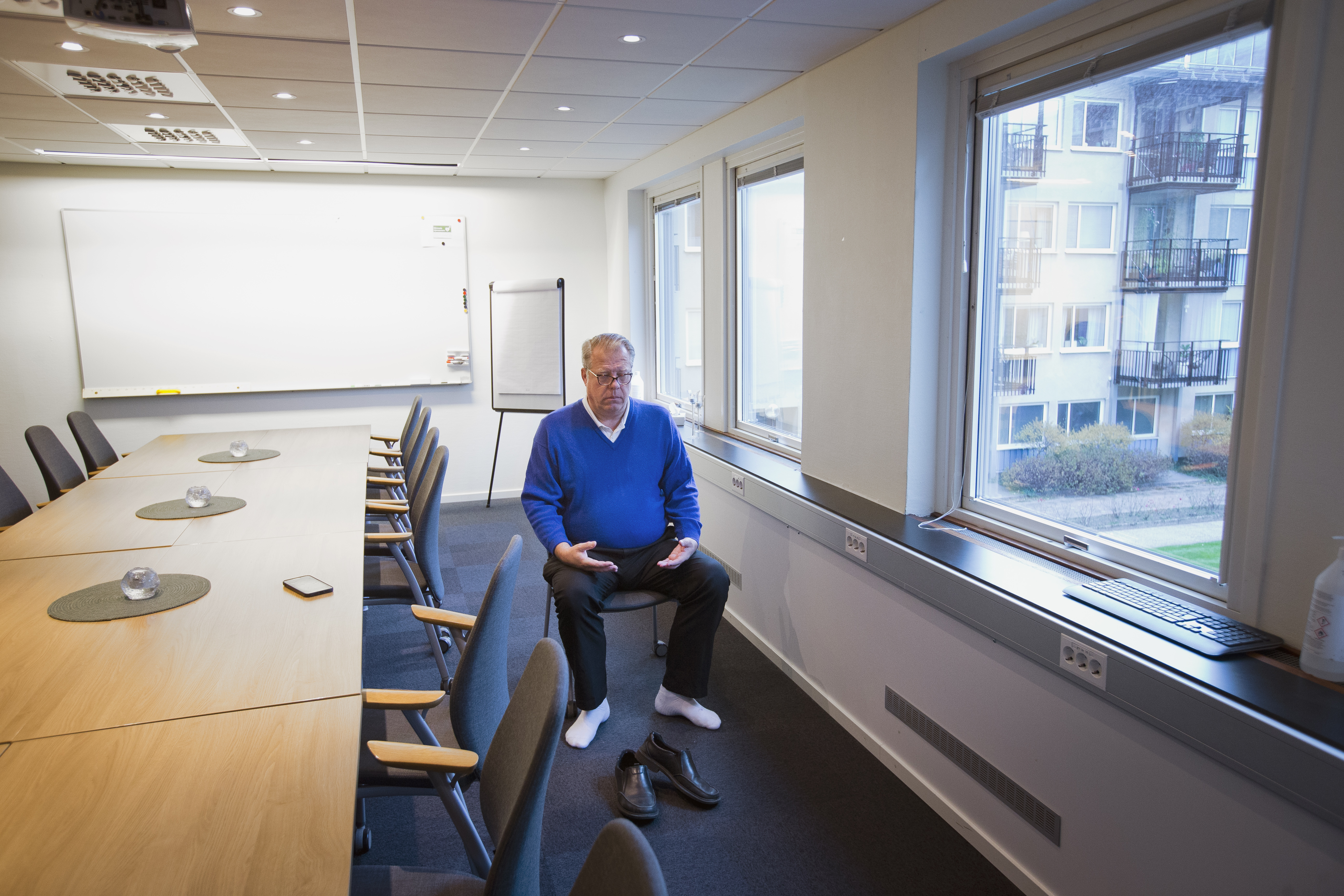 En man i blå tröja sitter på en stol i ett konferensrum utan skor, håller en telefon, nära stora fönster mot lägenheter.