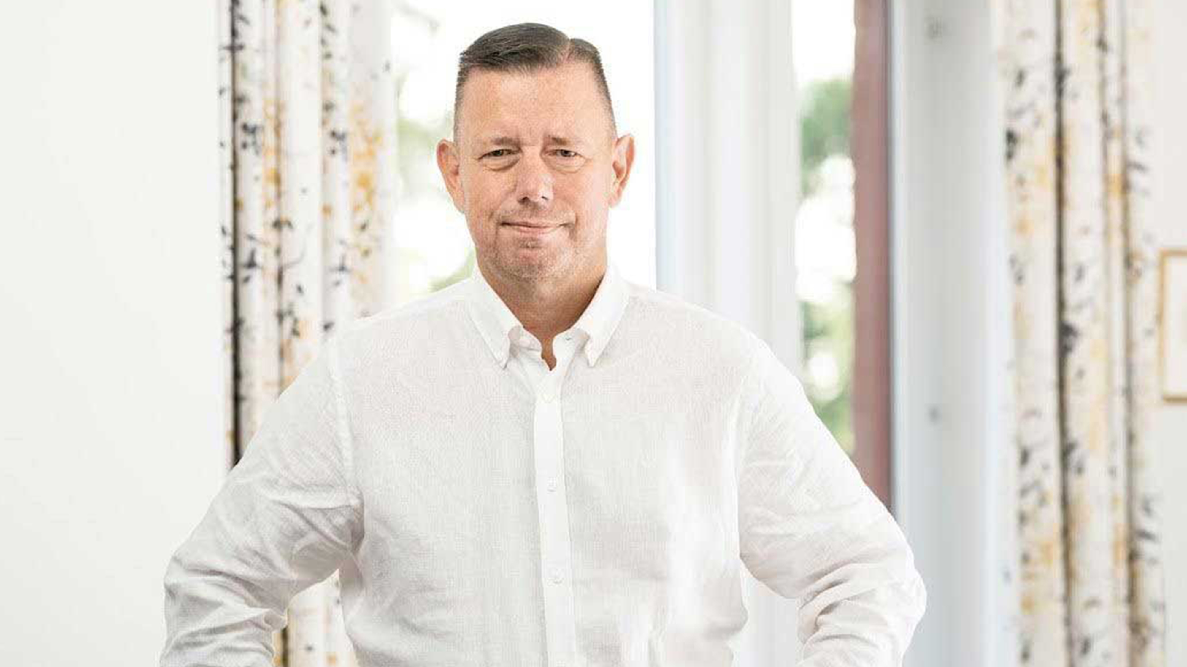 En man med vit skjorta står inomhus framför ett fönster med mönstrade gardiner och tittar mot kameran.