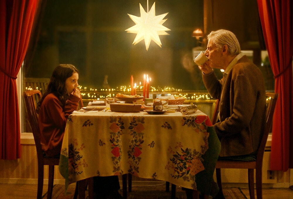 En äldre man och en ung flicka (skådespelarna Luiza Idrizi och Staffan Göthe) sitter mittemot varandra vid ett middagsbord med ljus, mat och en adventsstjärna över sig.