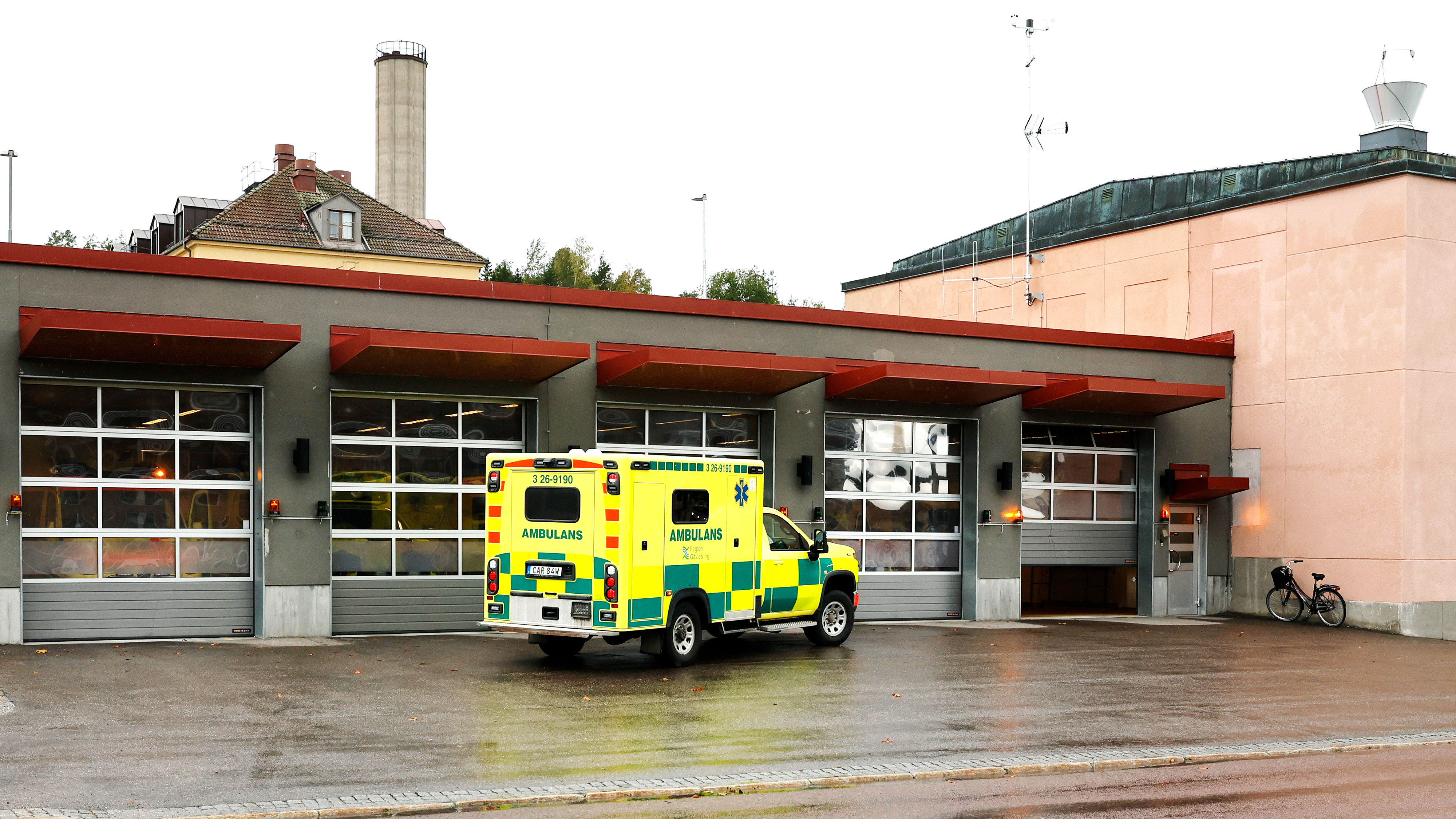 En gul och grön ambulans står parkerad utanför en byggnad med flera garageportar en regnig dag.