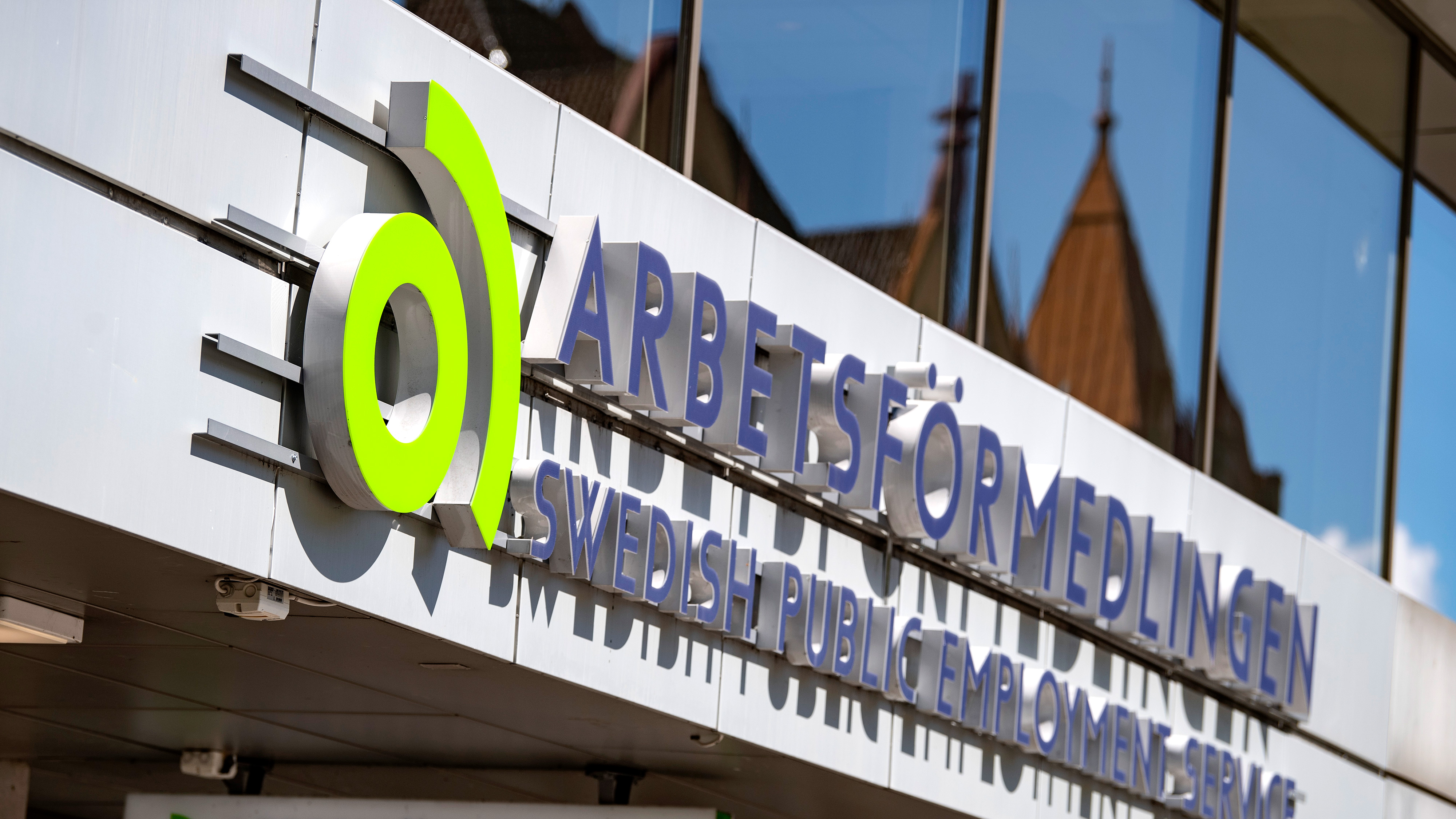 Skylt för Arbetsförmedlingen, Sveriges arbetsförmedling, monterad på en byggnadsfasad med speglingar i glasfönstren ovanförför.