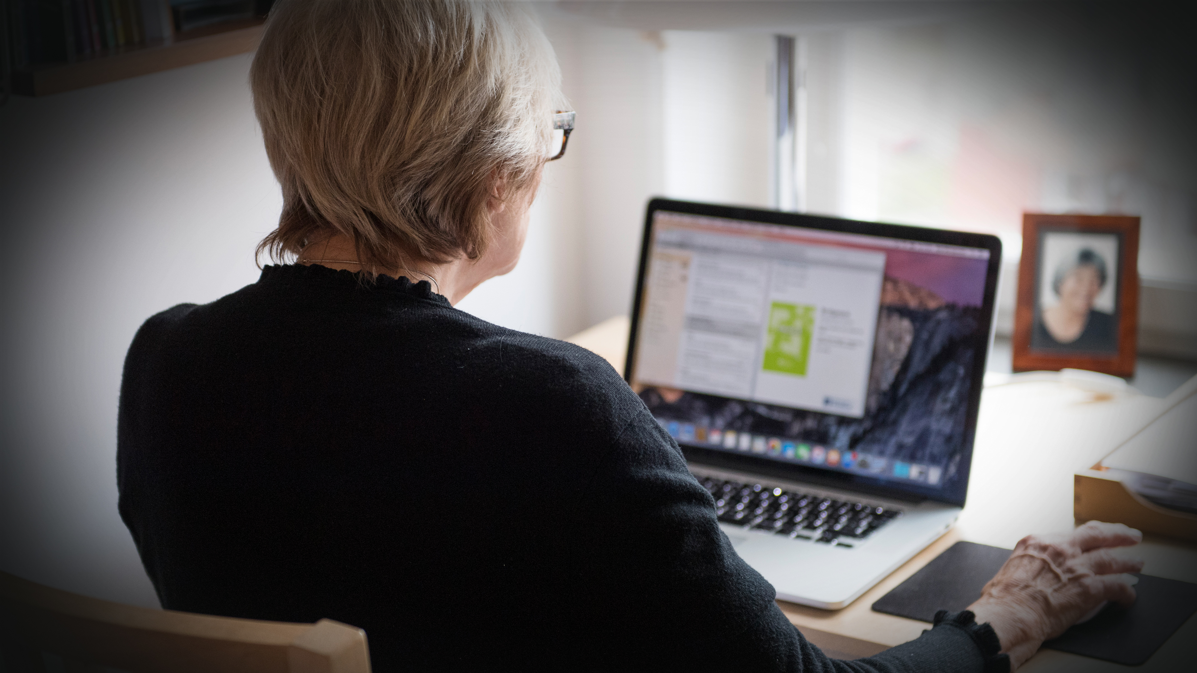 En äldre kvinna med kort grått hår sitter vid ett skrivbord och använder en bärbar dator, med foto, lampa och kontorssaker intill.