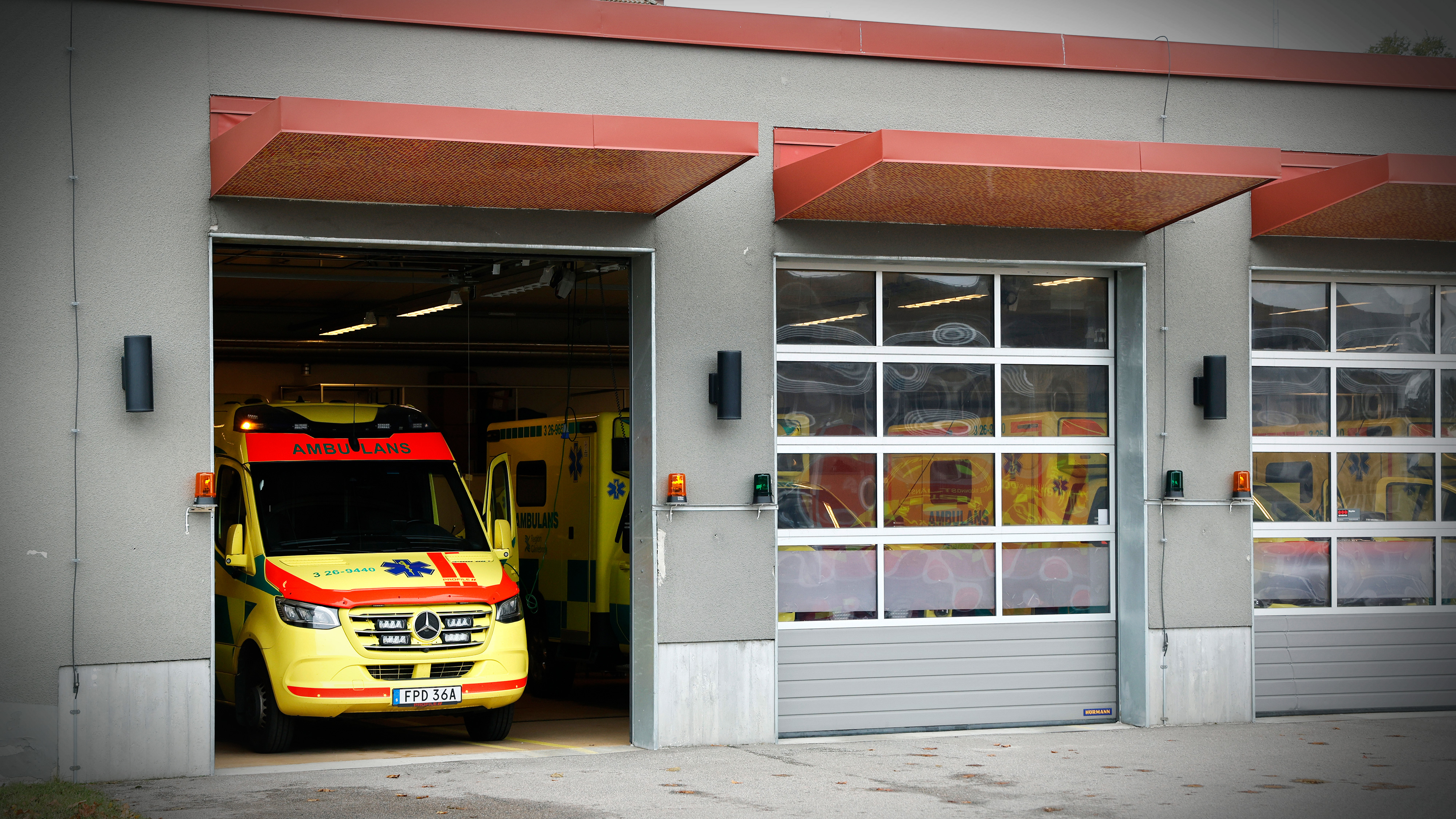 En ambulans står delvis parkerad utanför en garagebyggnad med två öppna och en stängd port vid en utryckningsstation.