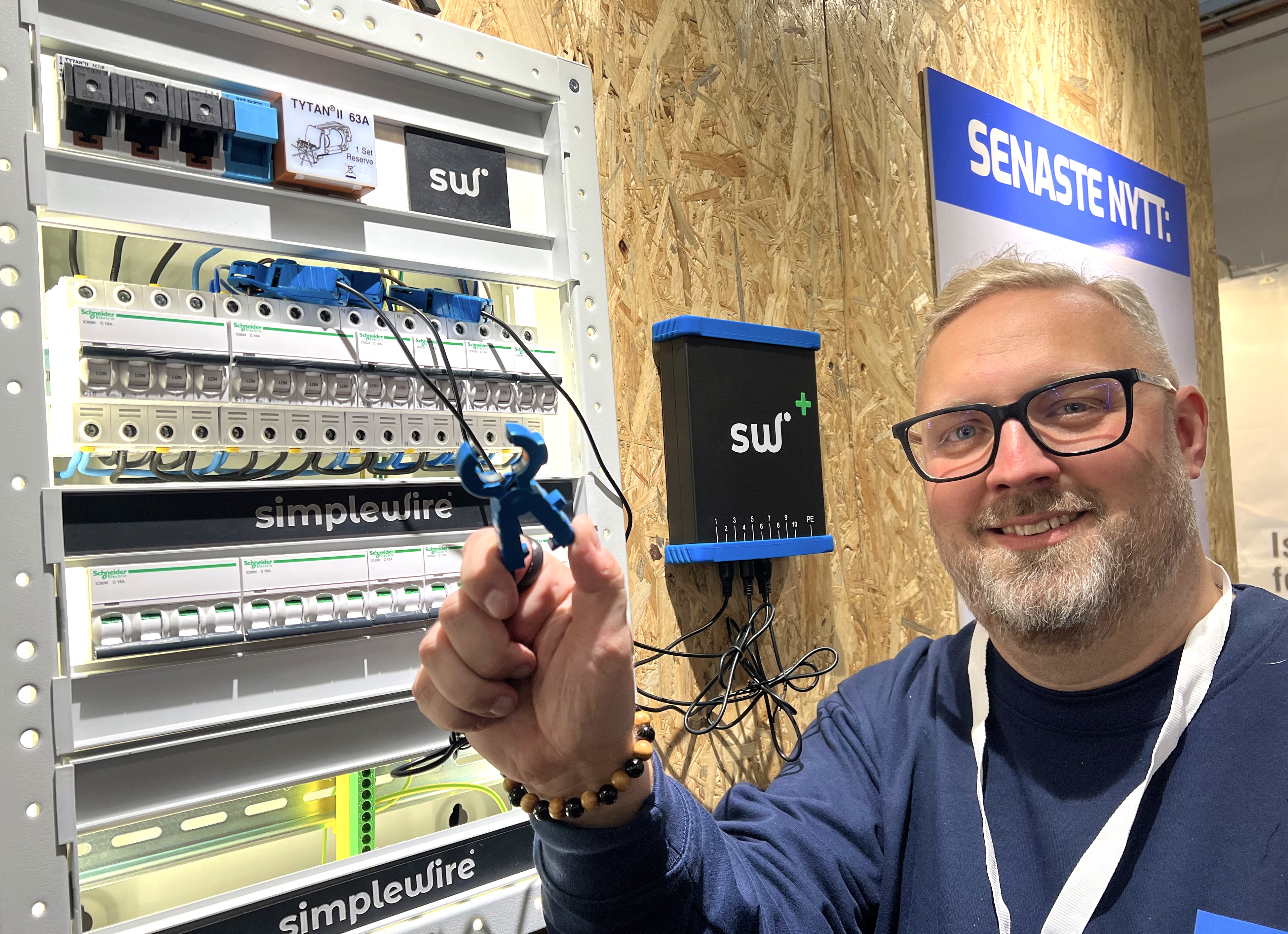 En man står bredvid ett öppet elskåp, håller ett blått verktyg och tittar mot kameran. Olika komponenter syns i skåpet.