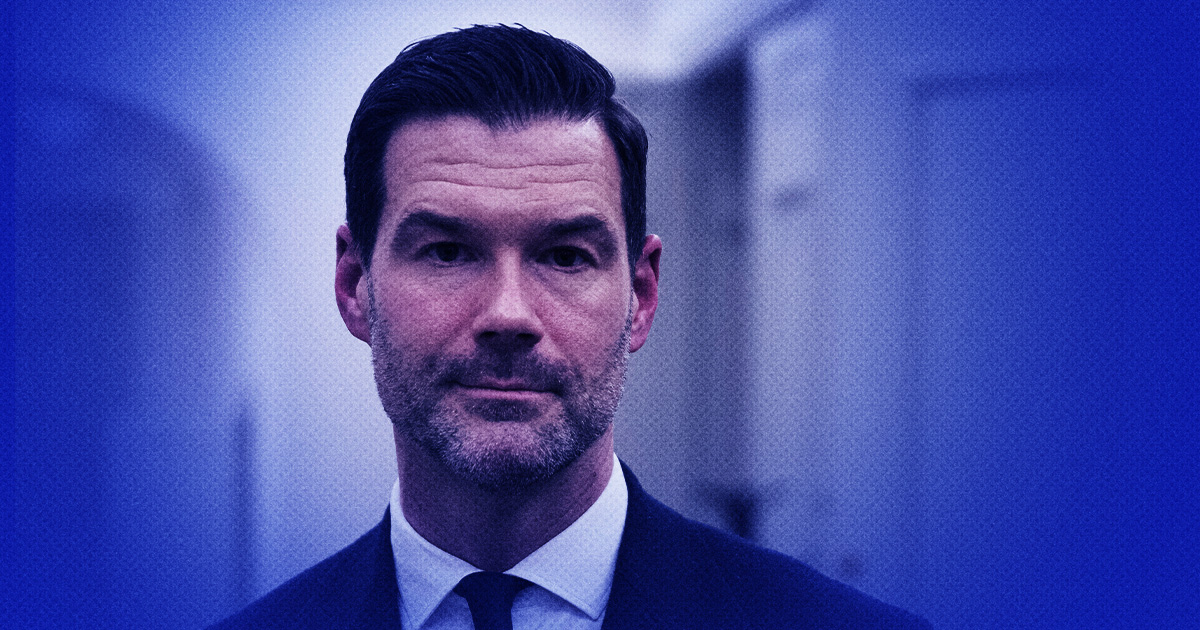 Johan Forsell med mörkt hår och skägg, iförd kostym och slipsar, står inomhus mot en oskarp blå bakgrund.