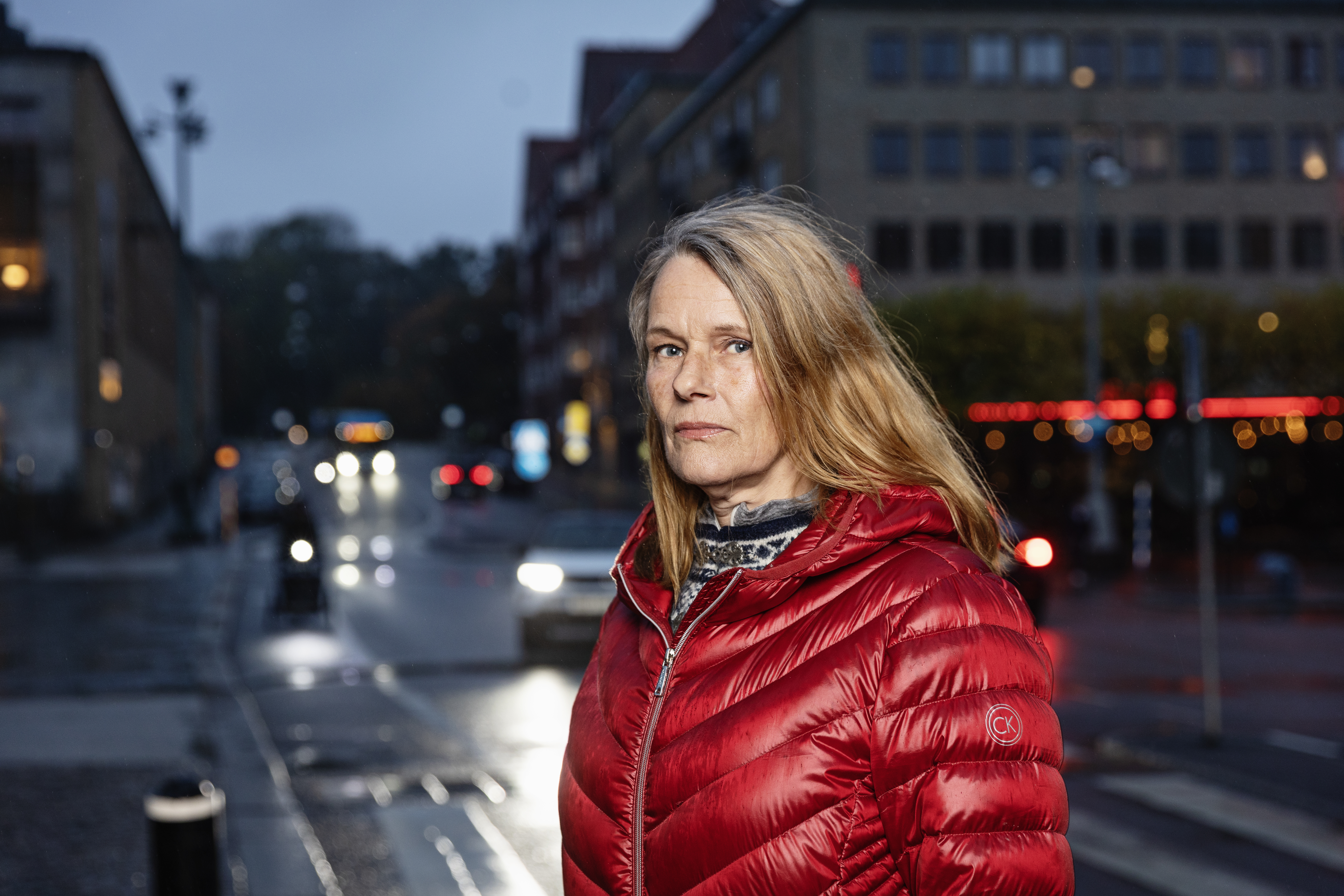 En kvinna med en röd dunjacka står på en stadsgata i skymningen, med bilar och byggnader i bakgrunden.
