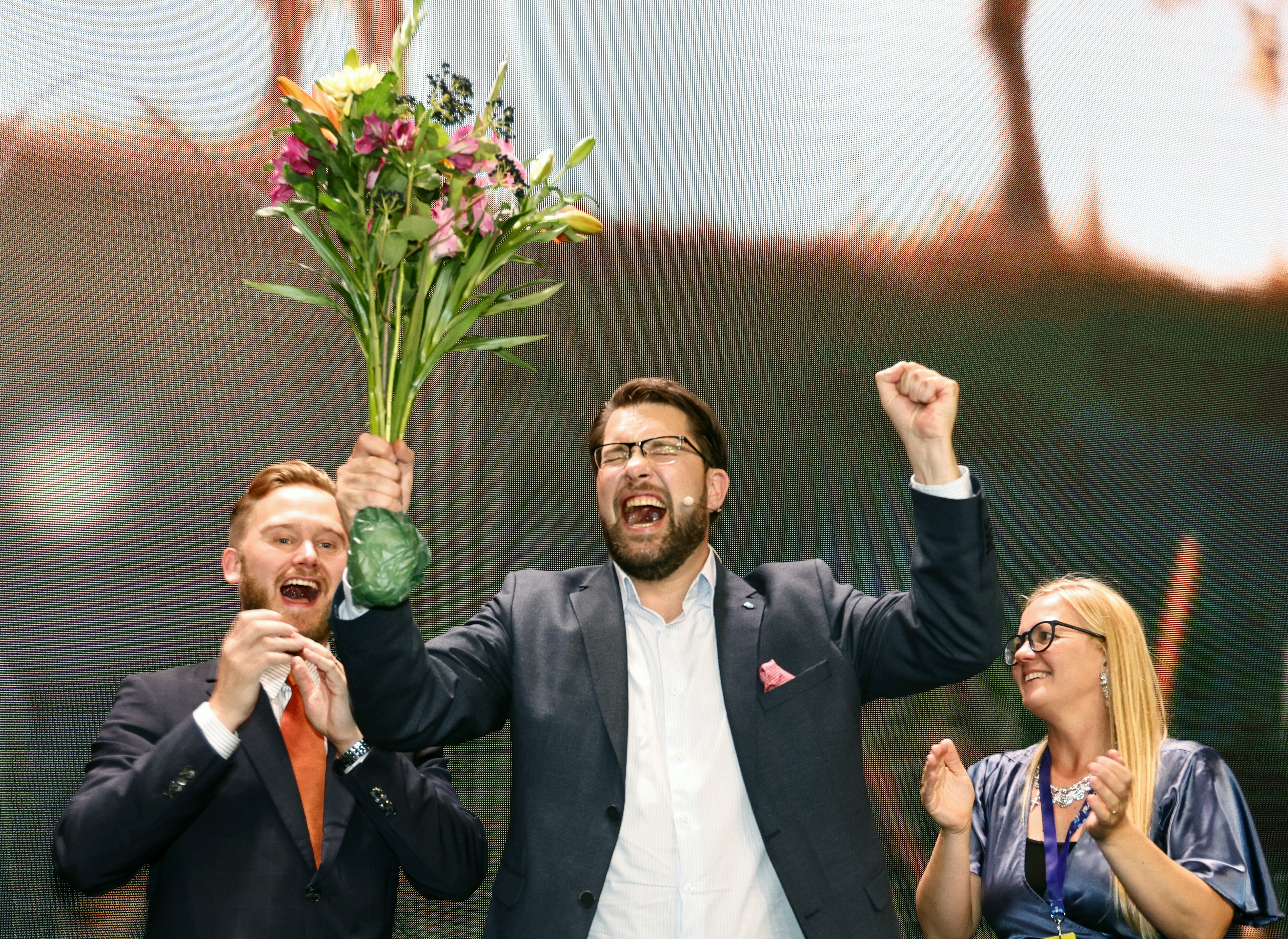 En man i kostym lyfter en bukett blommor och jublar, medan två personer bredvid honom ler och applåderar på scenen.