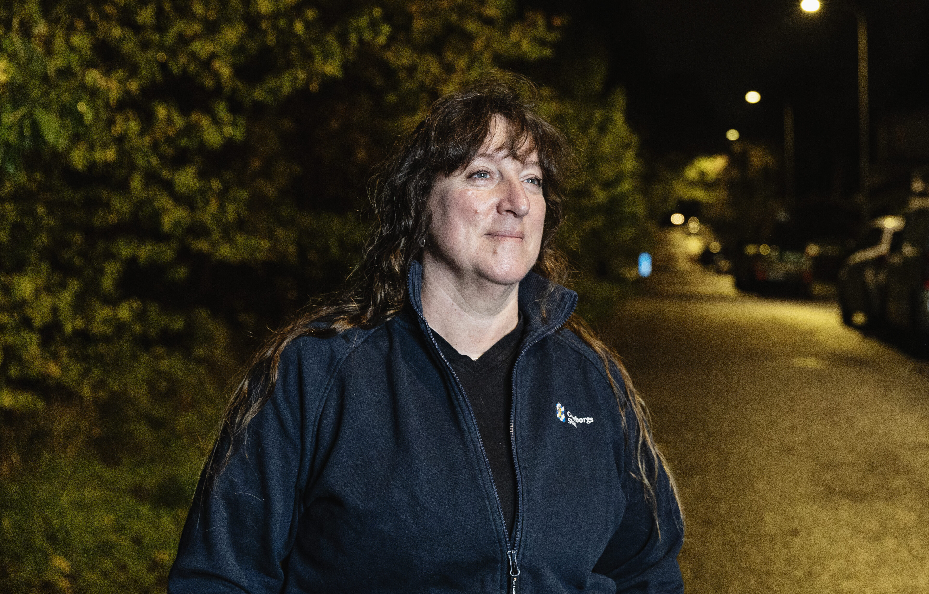 En kvinna med långt hår och en marinblå jacka står på en gata på natten, med träd och parkerade bilar i bakgrunden.