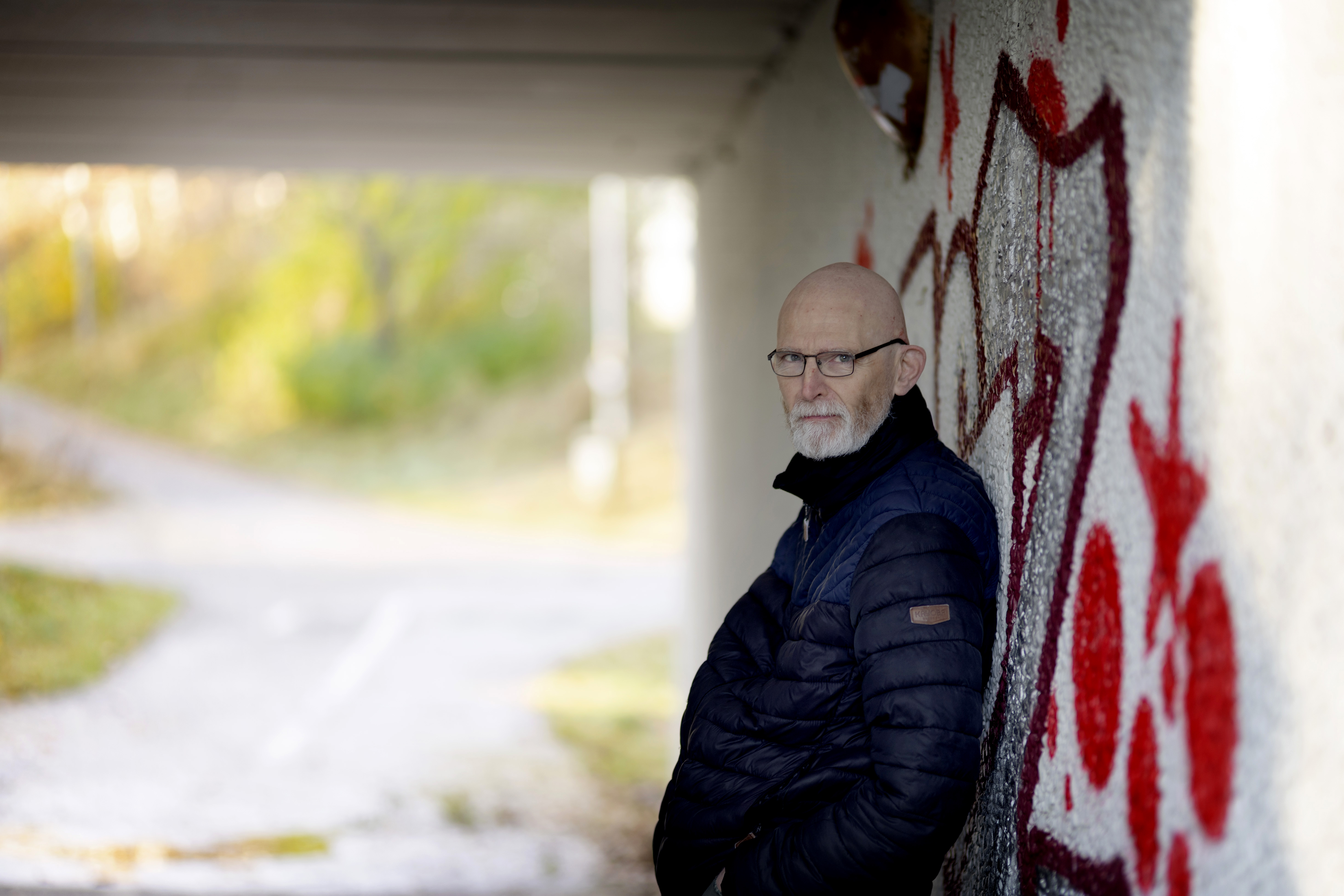 En man med glasögon och marinblå jacka lutar sig mot en vägg med graffiti under en viadukt och tittar mot kameran.