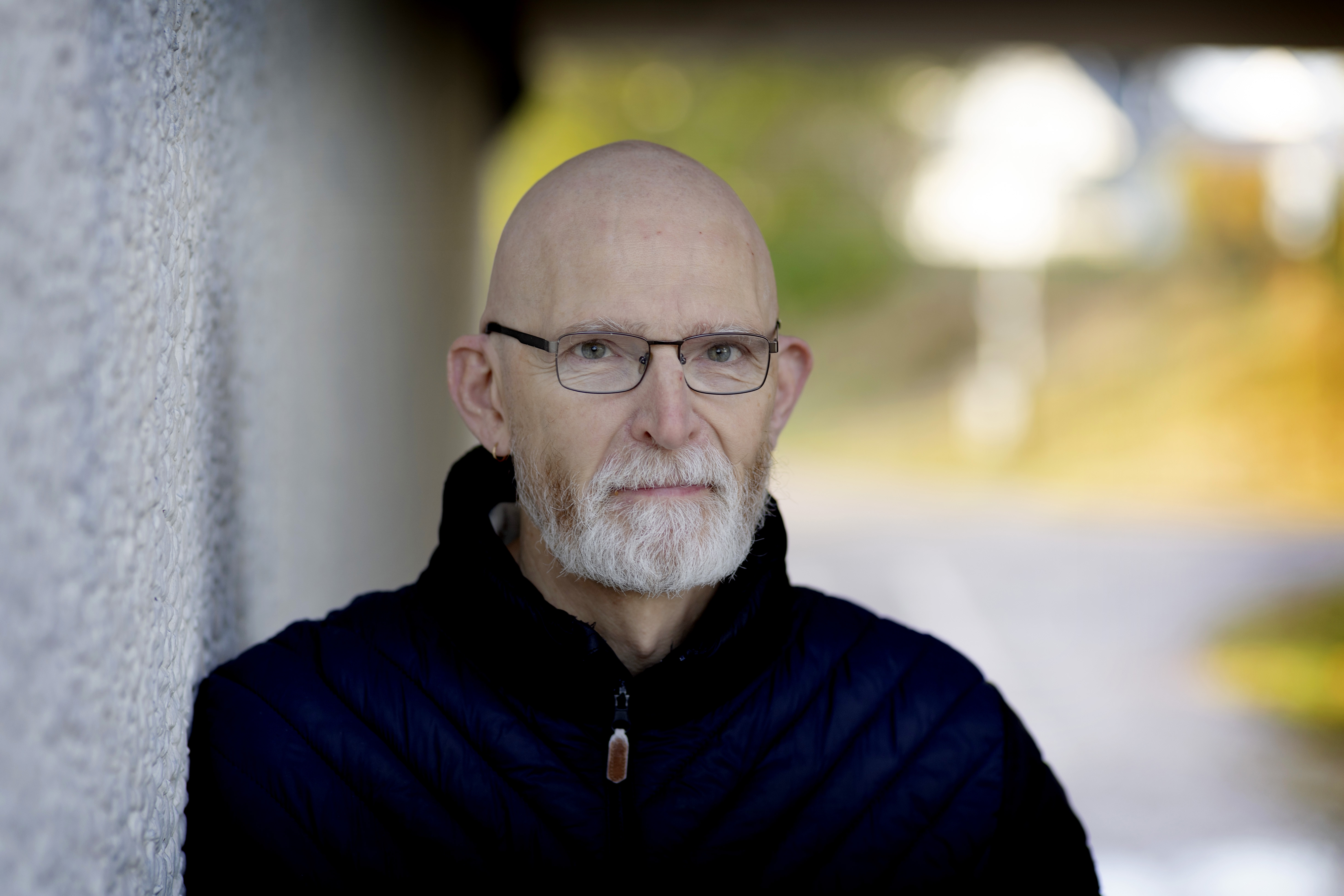 En man med glasögon och grått skägg står utomhus lutad mot en vit vägg. I bakgrunden syns gata och grönska.