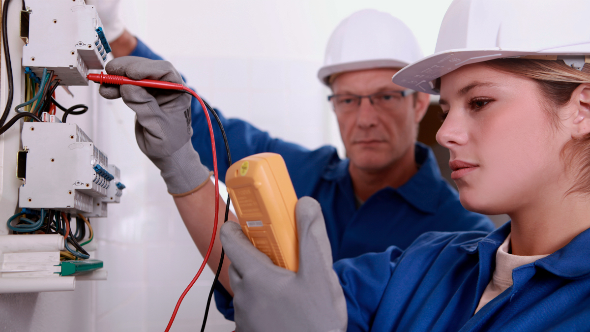 Två elektriker med skyddsutrustning testar en elcentral med en multimeter. En håller apparaten, den andra justerar proberna.