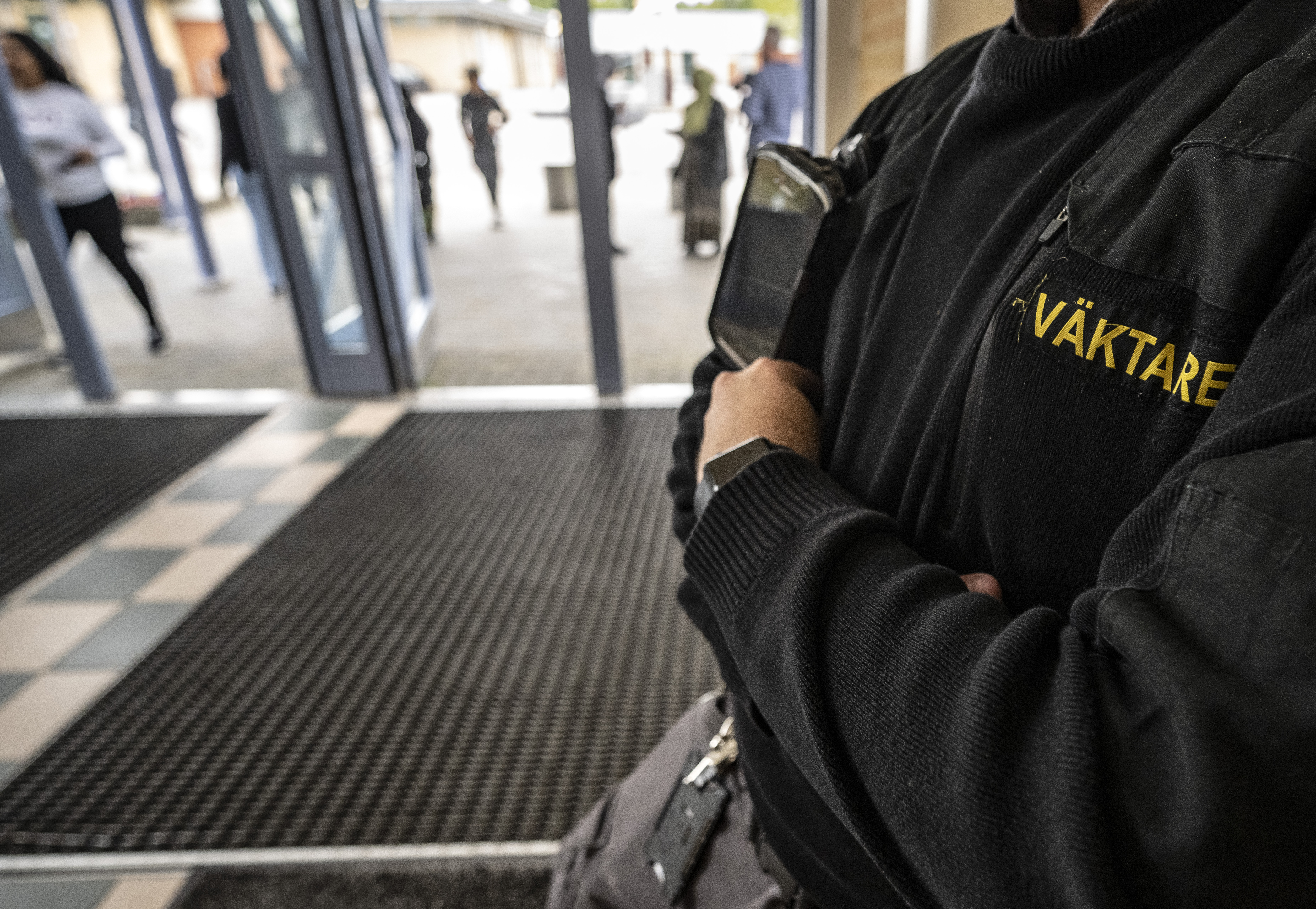 En person i svart uniform med texten "Väktare" står med korslagda armar vid en ingång, personer finns i bakgrunden.