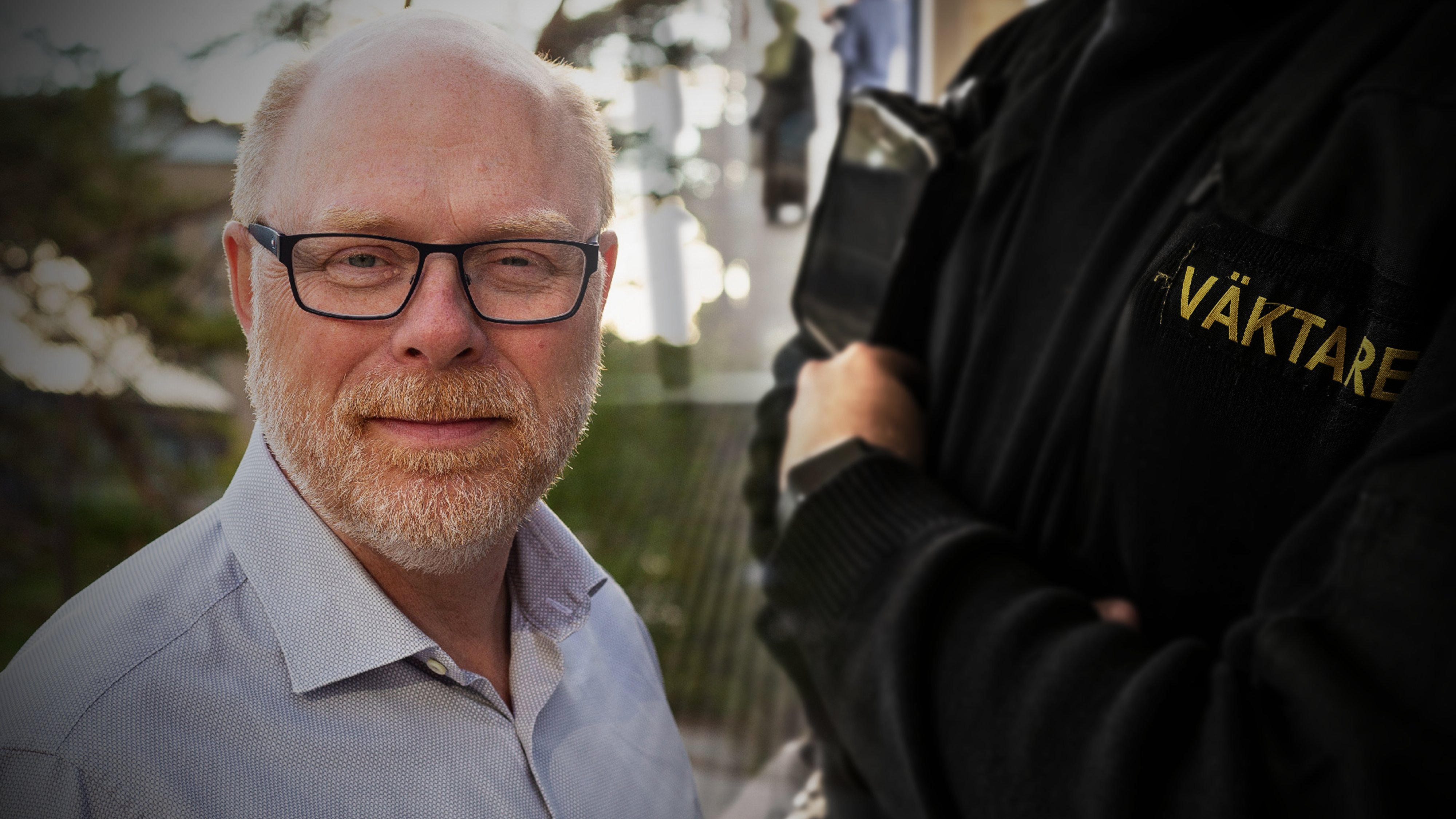 En man med glasögon och skägg står utomhus, bredvid honom en person i svarta kläder med texten "VÄKTARE" på ärmen.