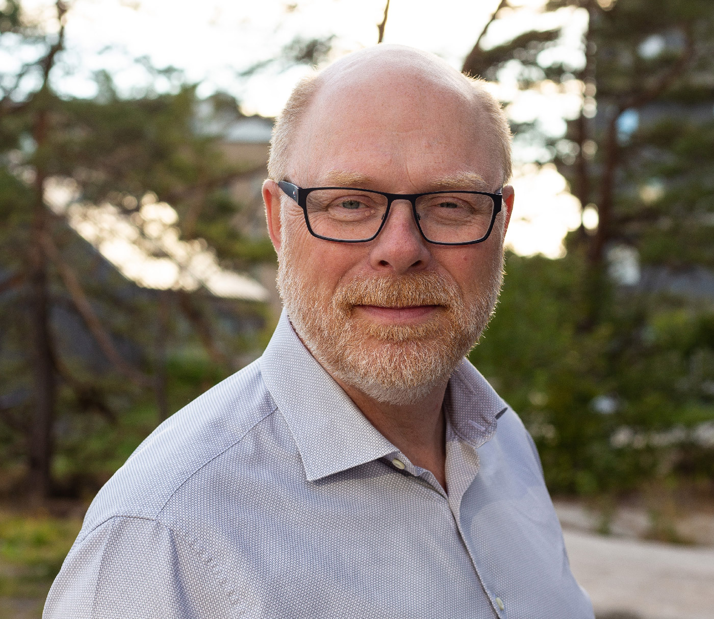En medelålders man med glasögon, skägg och ljust hår står utomhus i ljus skjorta. Träd och byggnader syns i bakgrunden.