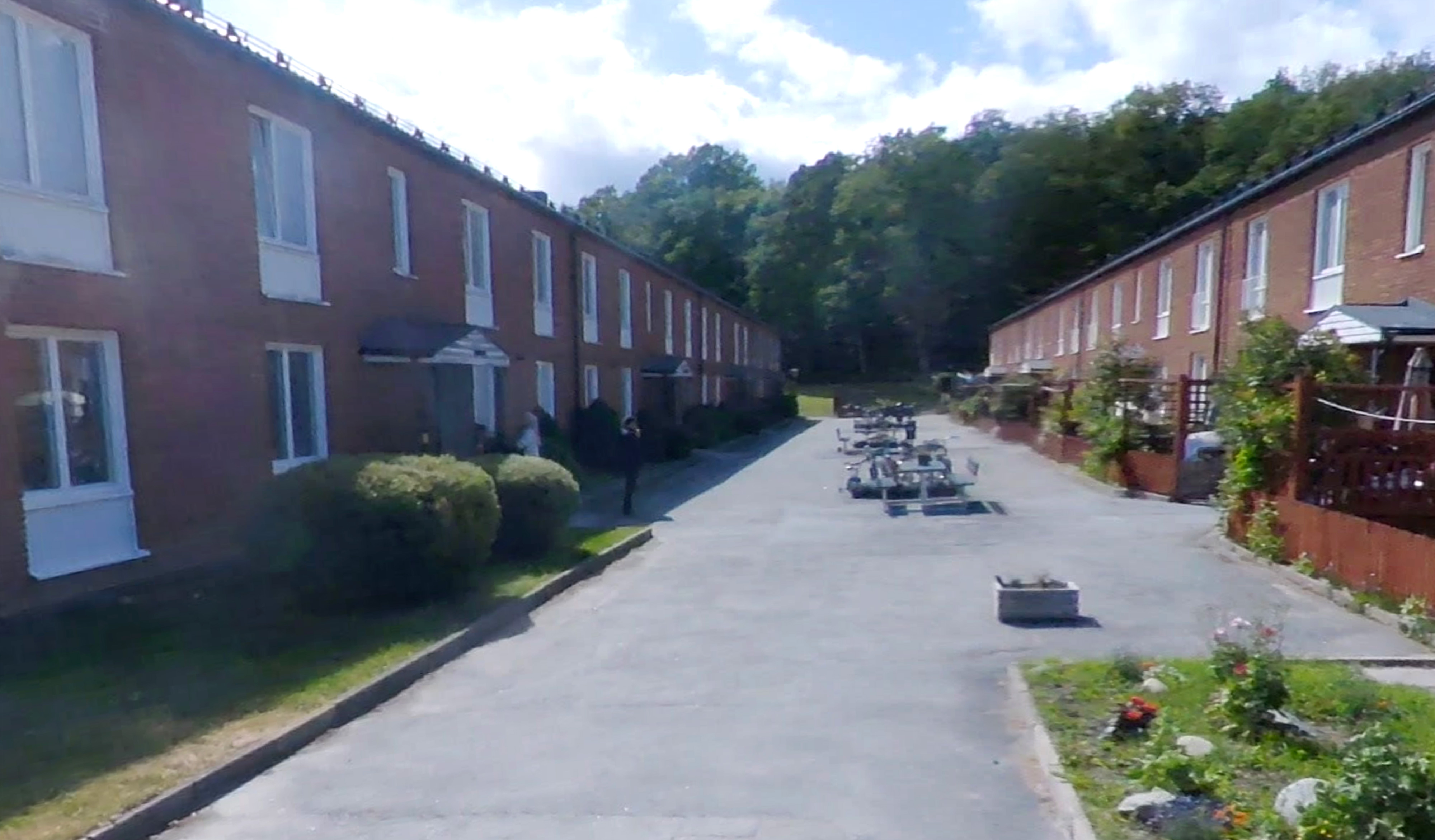 En bostadsgata med rader av tegelradhus på båda sidor, mittgång med bänkar och planteringar, omgiven av växtlighet och träd.