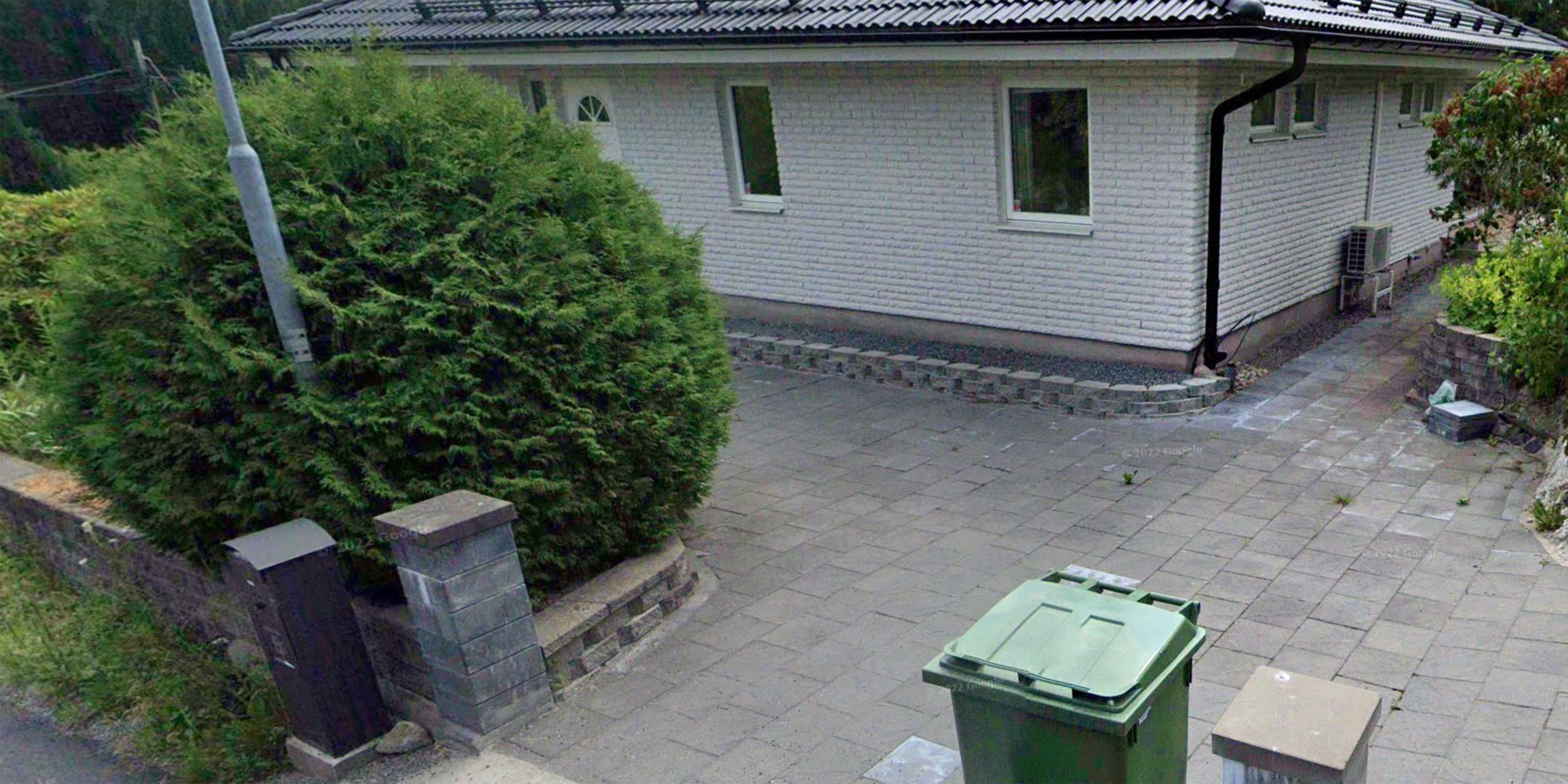 En stenlagd uppfart med en grön soptunna framför ett ljust tegelhus, intill en buske och en brevlåda vid ingången.