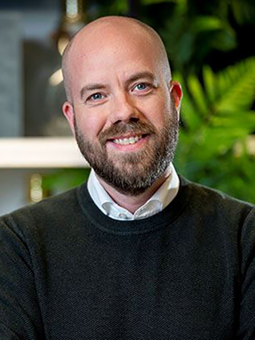 En man med rakat huvud och skägg, iförd mörk tröja över vit skjorta, ler mot kameran med grönska i bakgrunden.