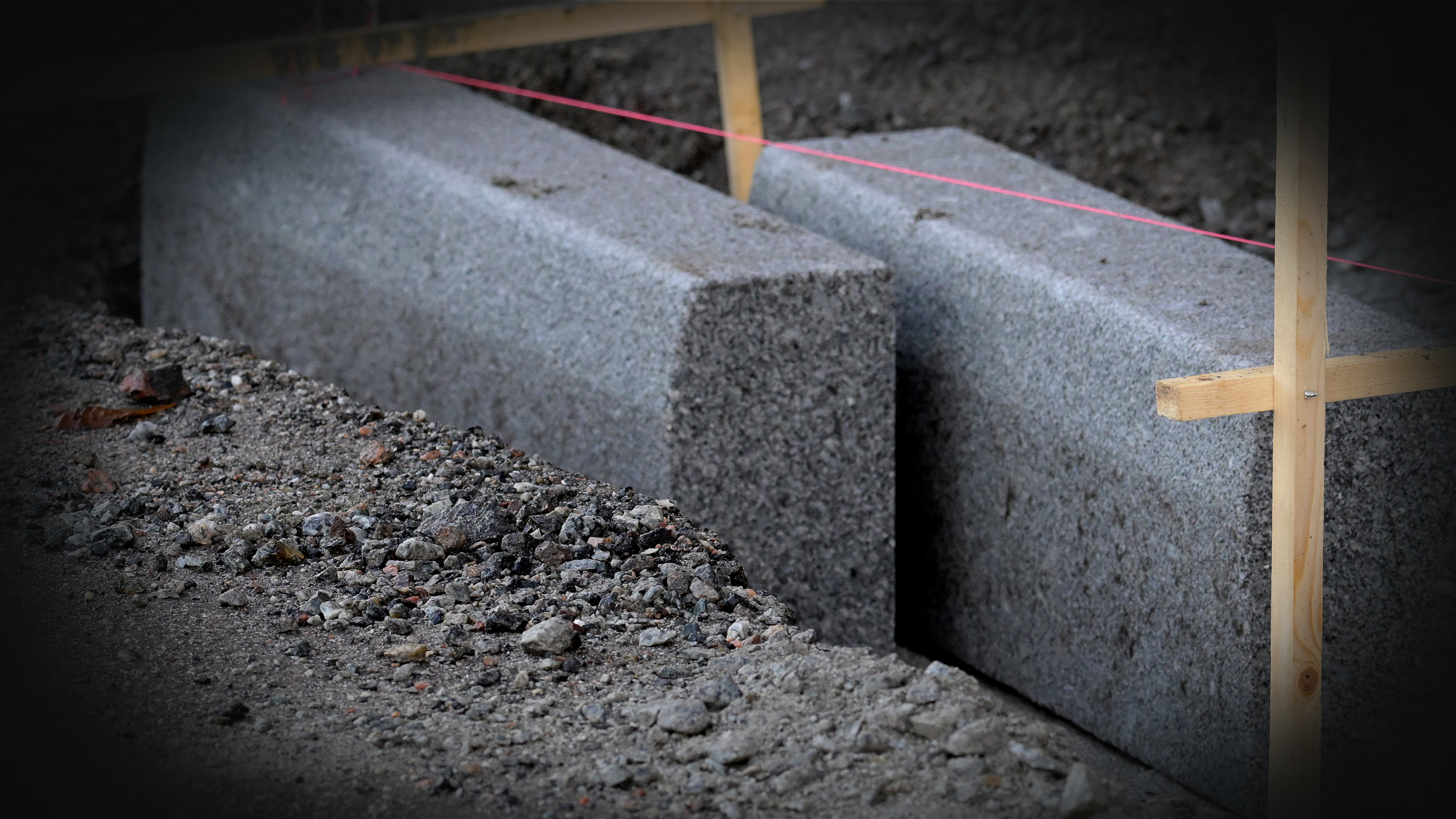 Två betongkantstenar installeras, utlinjerade med rosa snöre och träguider, omgivna av jord och grus på en byggarbetsplats.