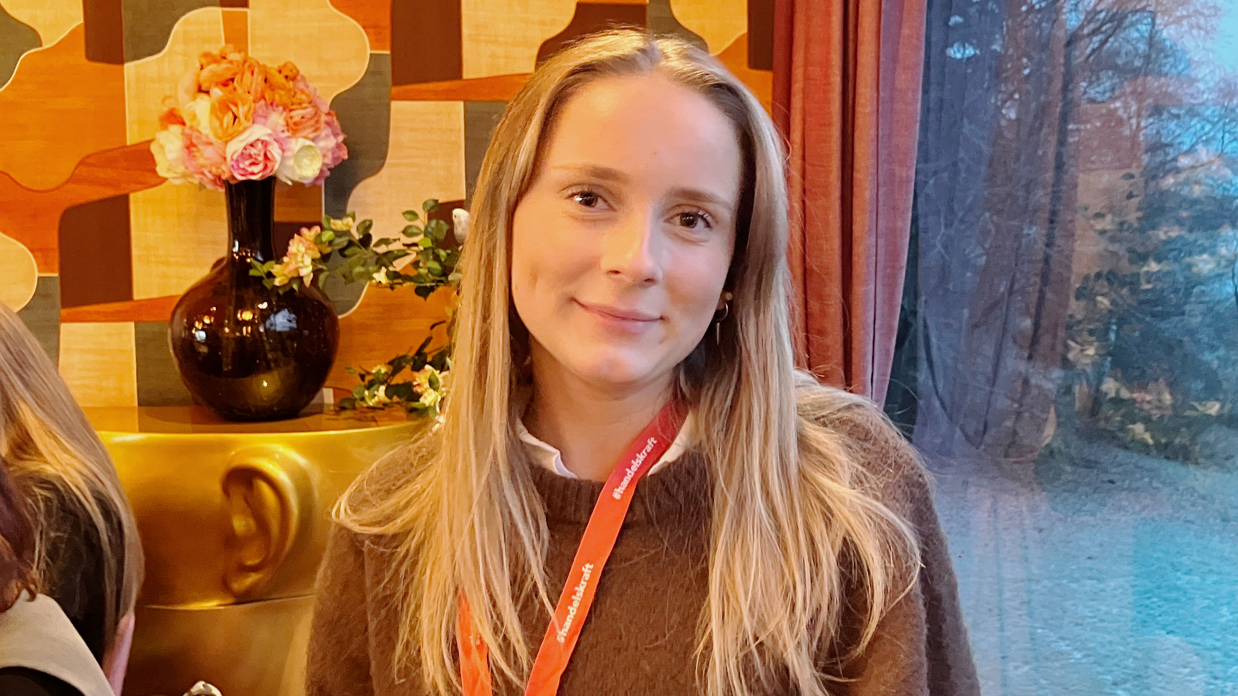 En kvinna med långt blont hår sitter inomhus. Hon har en brun tröja och ett rött band. En vas med blommor och mönstrad tapet syns bakom.