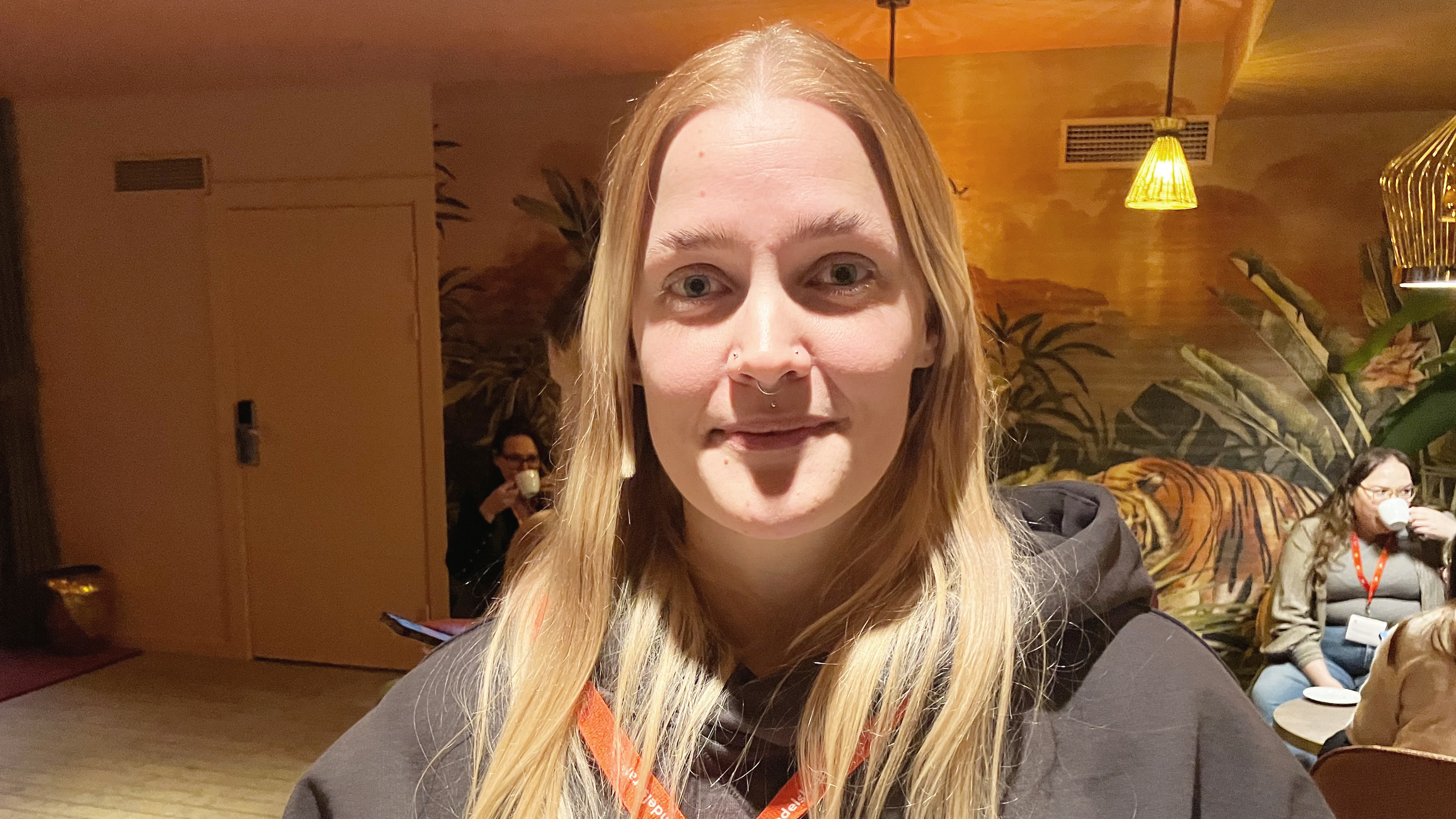 En kvinna med långt blont hår och näsring står inomhus med mörk huvtröja och rött nyckelband. Folk sitter, tiger på vägg.