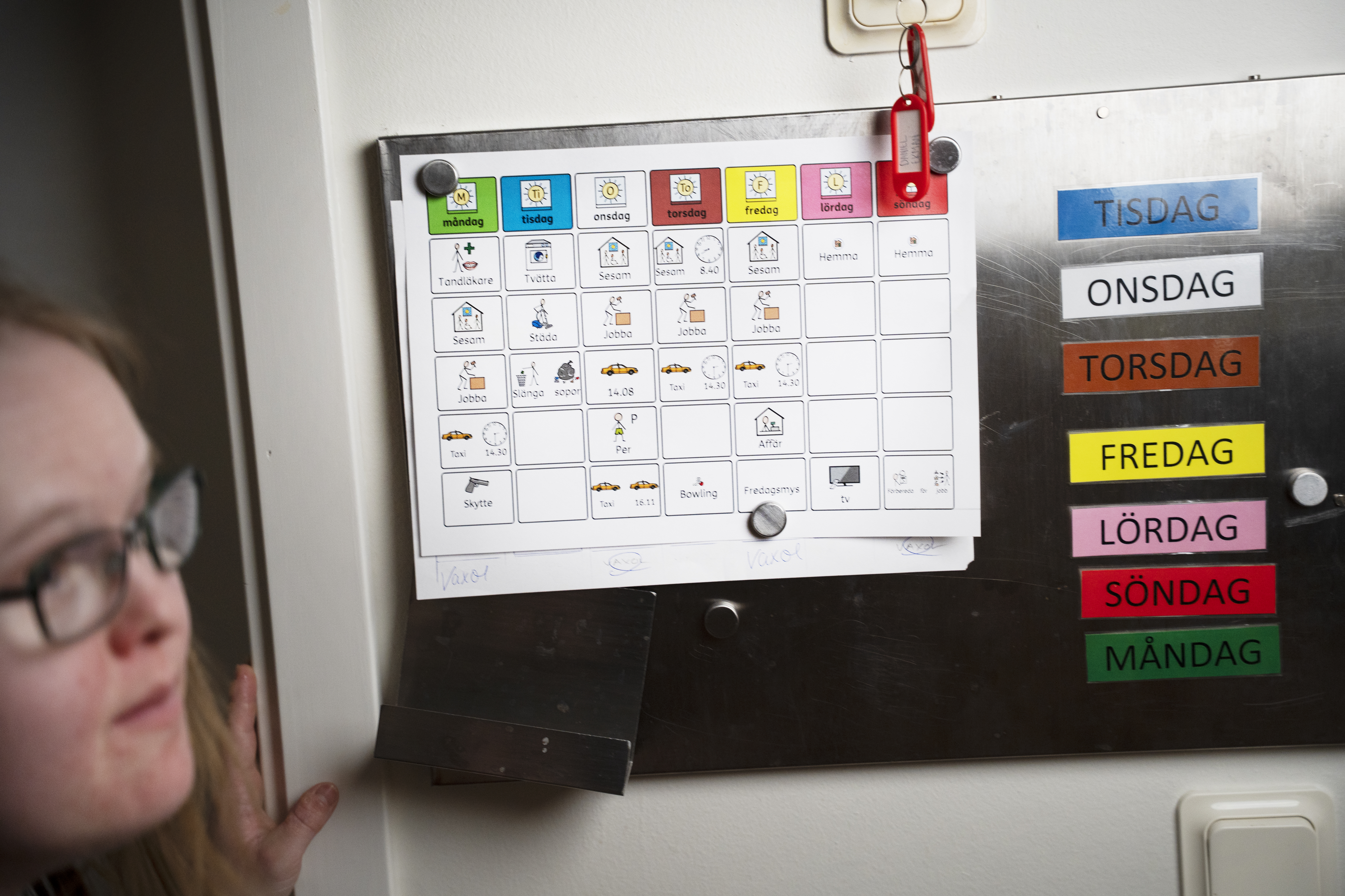 Mia med sitt veckoschema med symboler, som sitter på en metalltavla bredvid färgade etiketter för veckodagar på svenska.