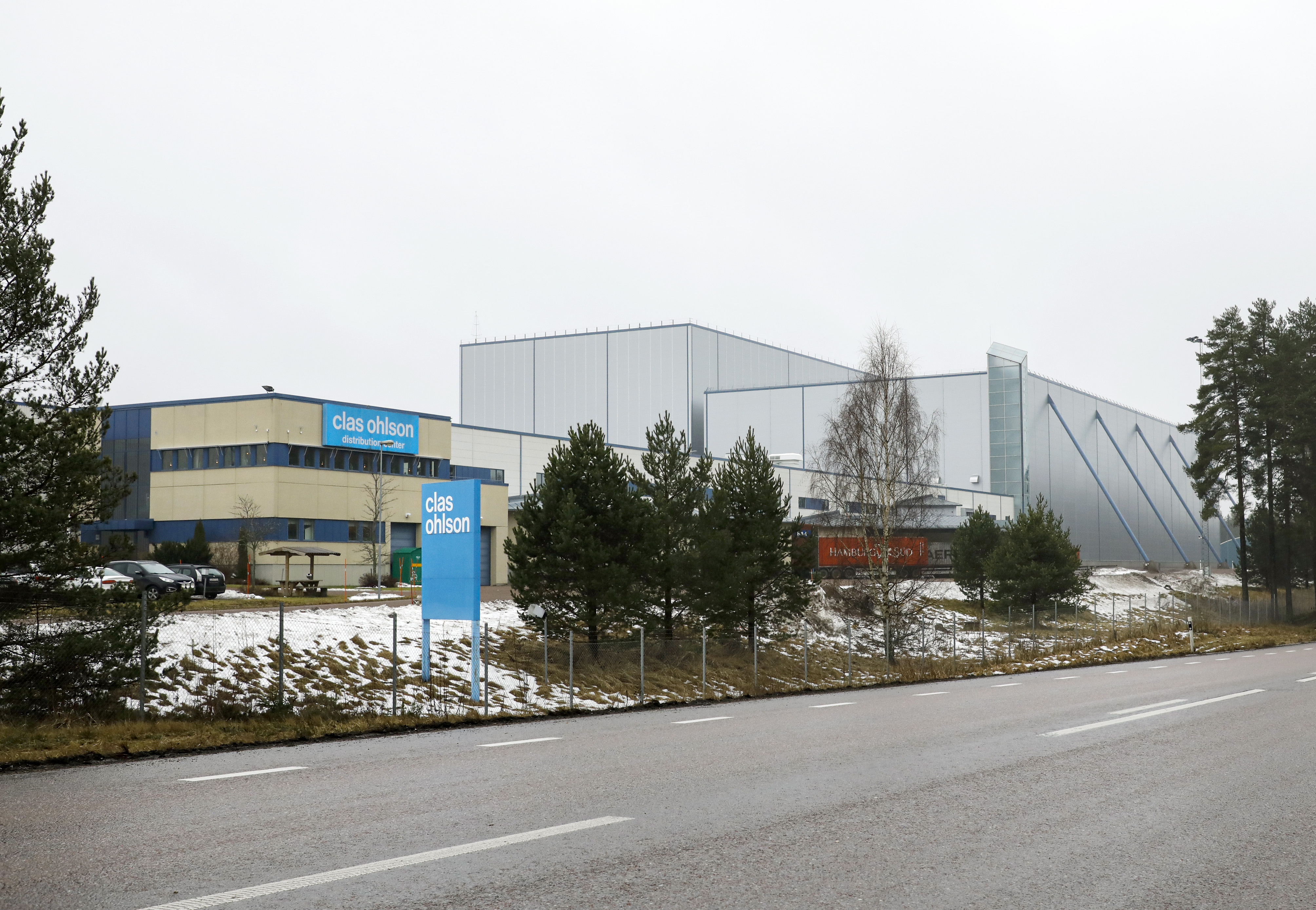 En stor lagerbyggnad med ”Clas Ohlson”-skyltar, omgiven av träd och lite snö, sedd från andra sidan vägen en mulen dag.