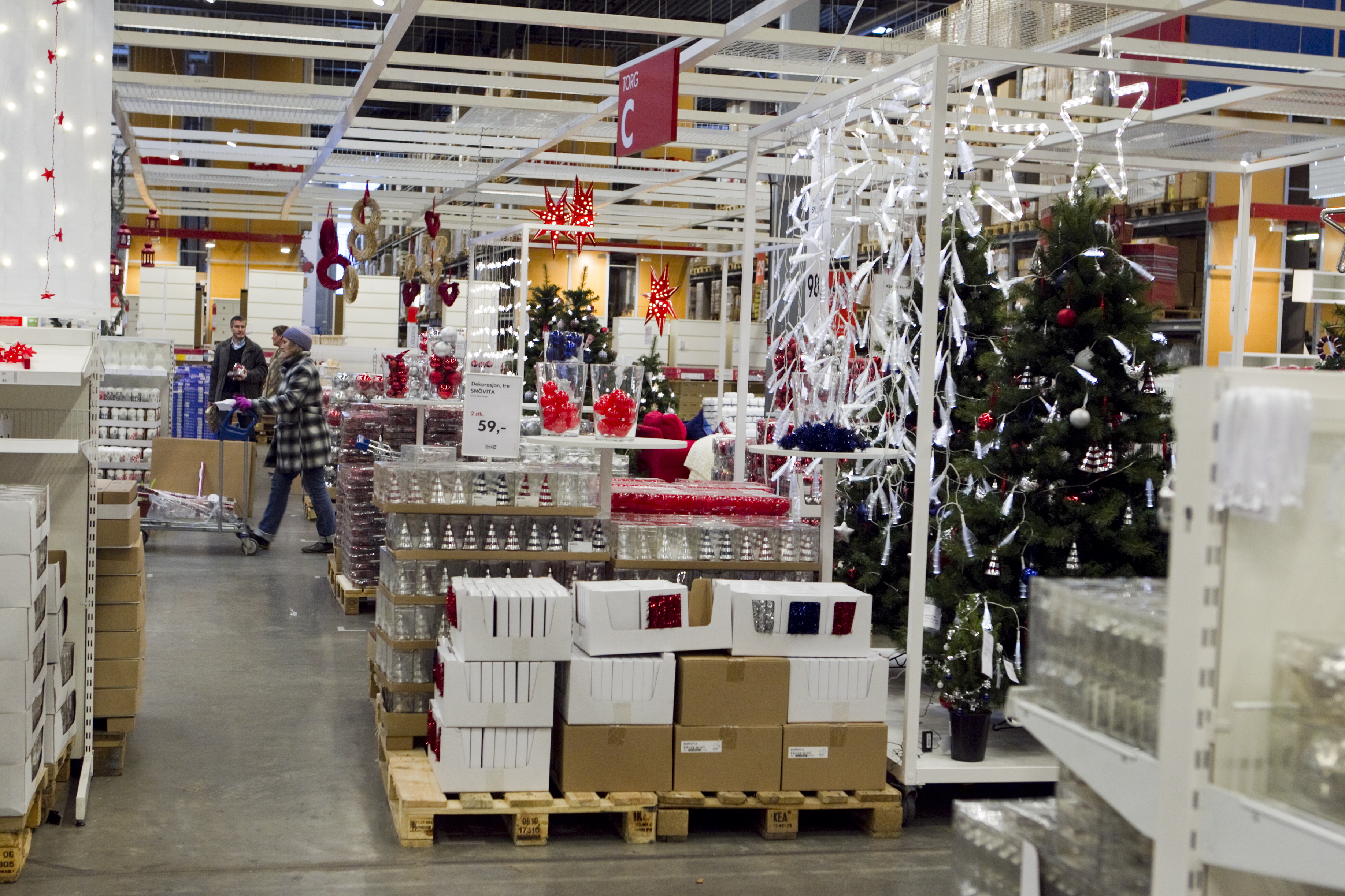 Butiksgång med juldekorationer, konstgjorda granar, prydnader och ljus. Kunder tittar på varorna på hyllor och pallar.