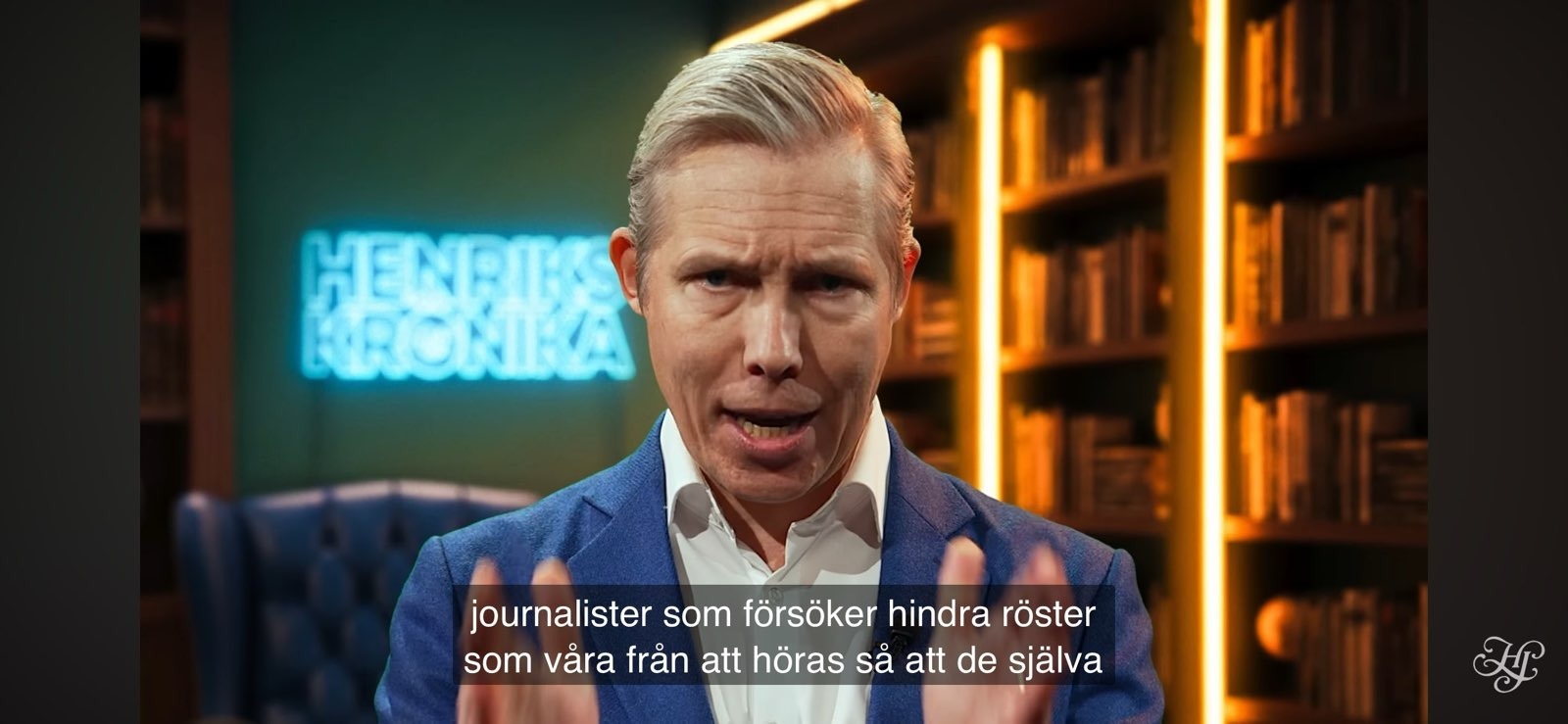 En man i blå kostym talar till kameran i ett bibliotek. Svenska undertexter visas längst ner på bilden.